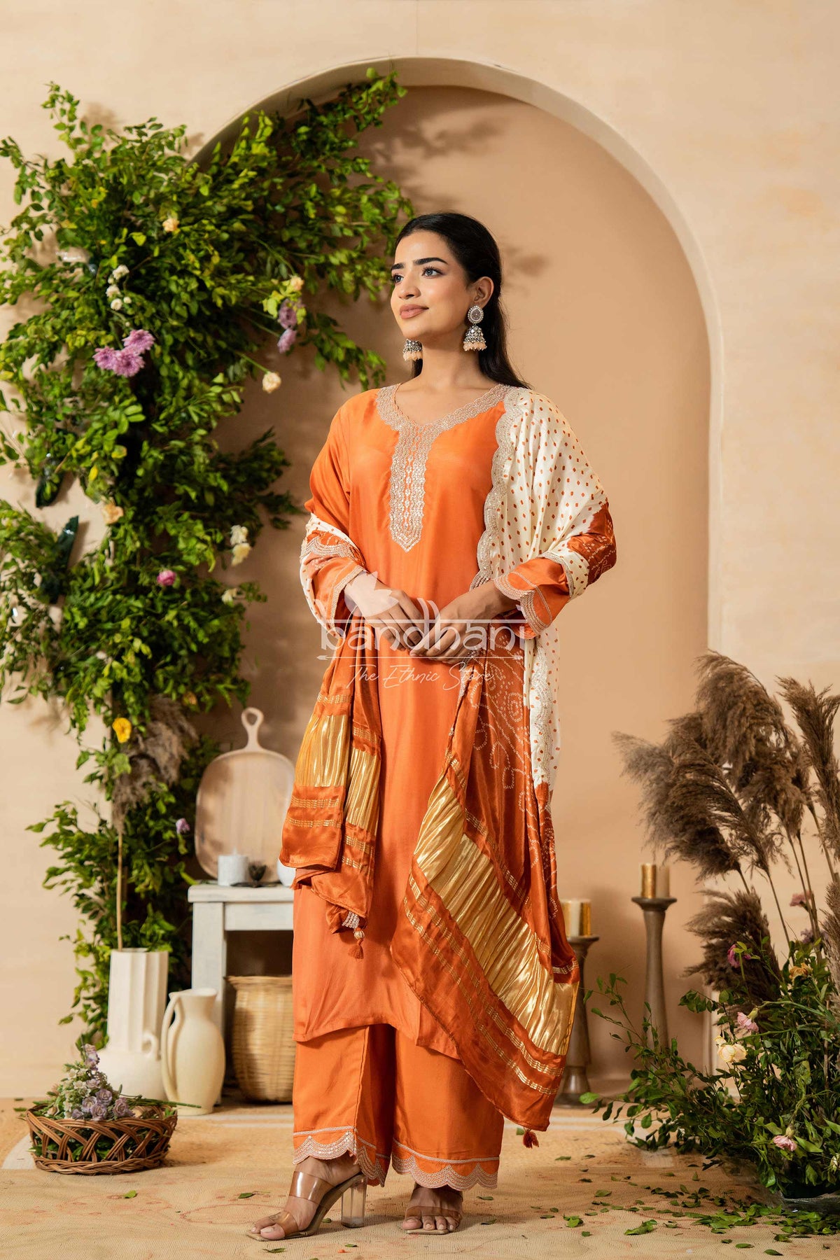 Woman in an orange and gold traditional outfit standing in a decorated indoor setting with plants and candles.
