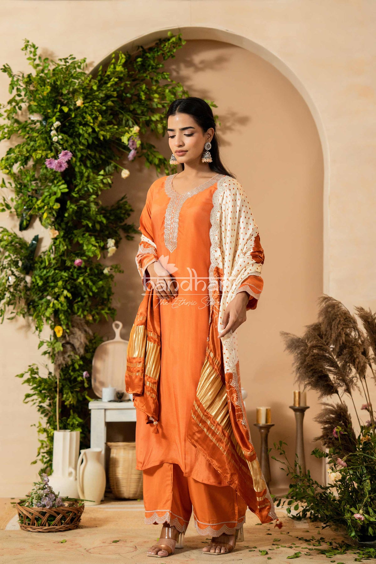 Woman in an orange traditional outfit standing in a decorated indoor setting with plants and candles.