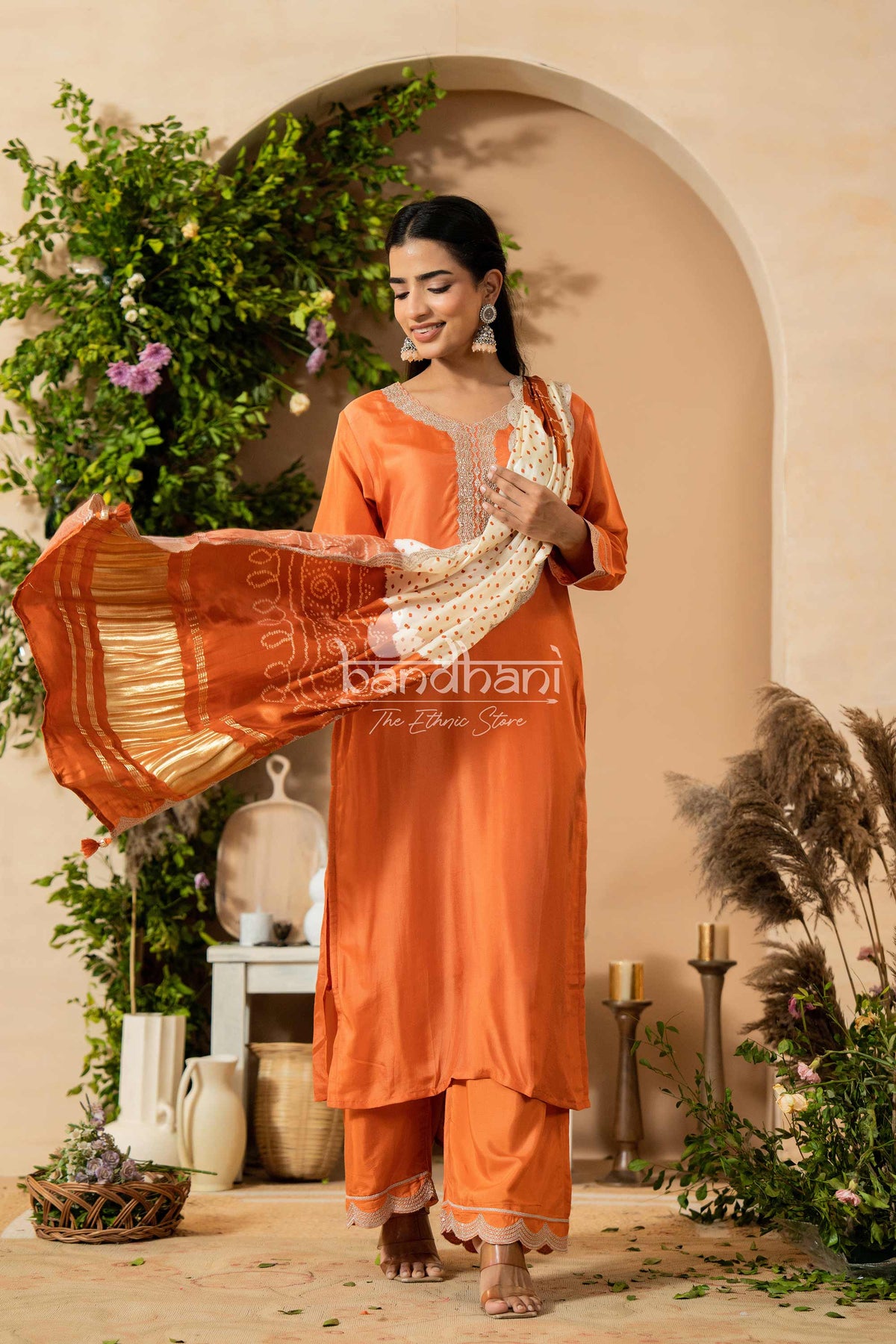 Woman in an orange traditional outfit with 'bandhani' text, standing in a decorated outdoor setting.