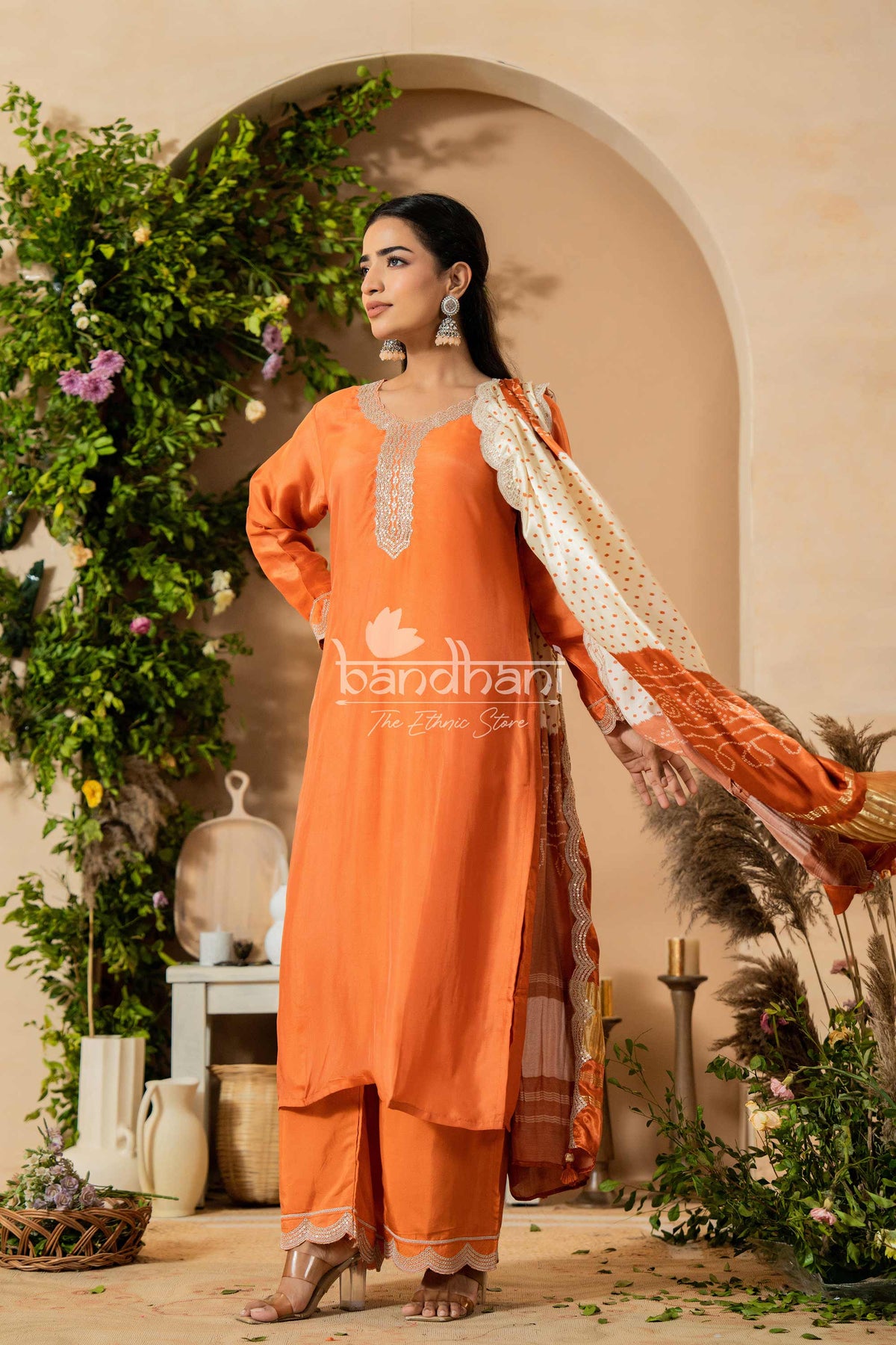 Woman in an orange traditional outfit with a white dupatta, standing in a decorated indoor setting with plants and decor.