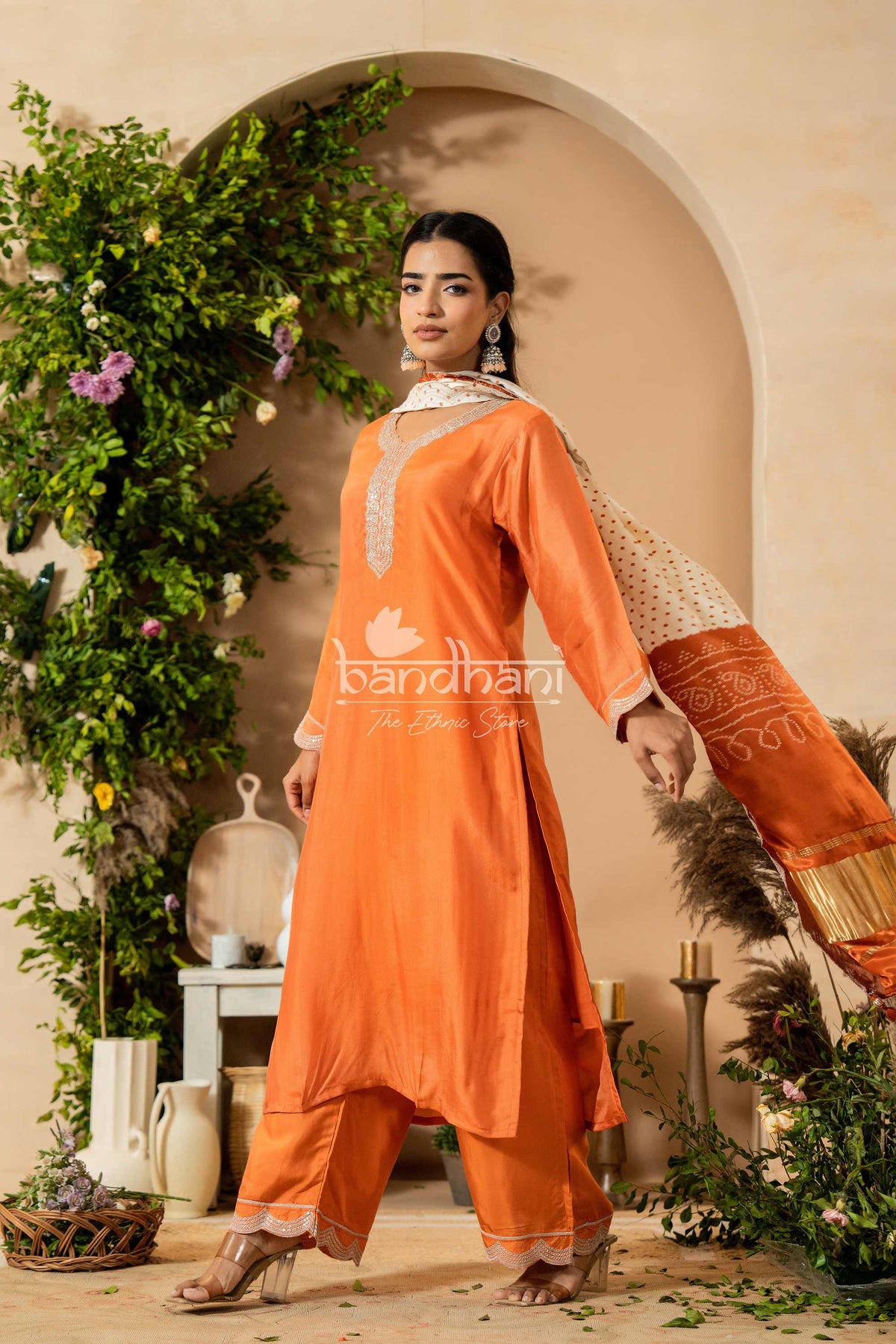 Woman in an orange traditional outfit standing in a decorative setting with plants and archway.