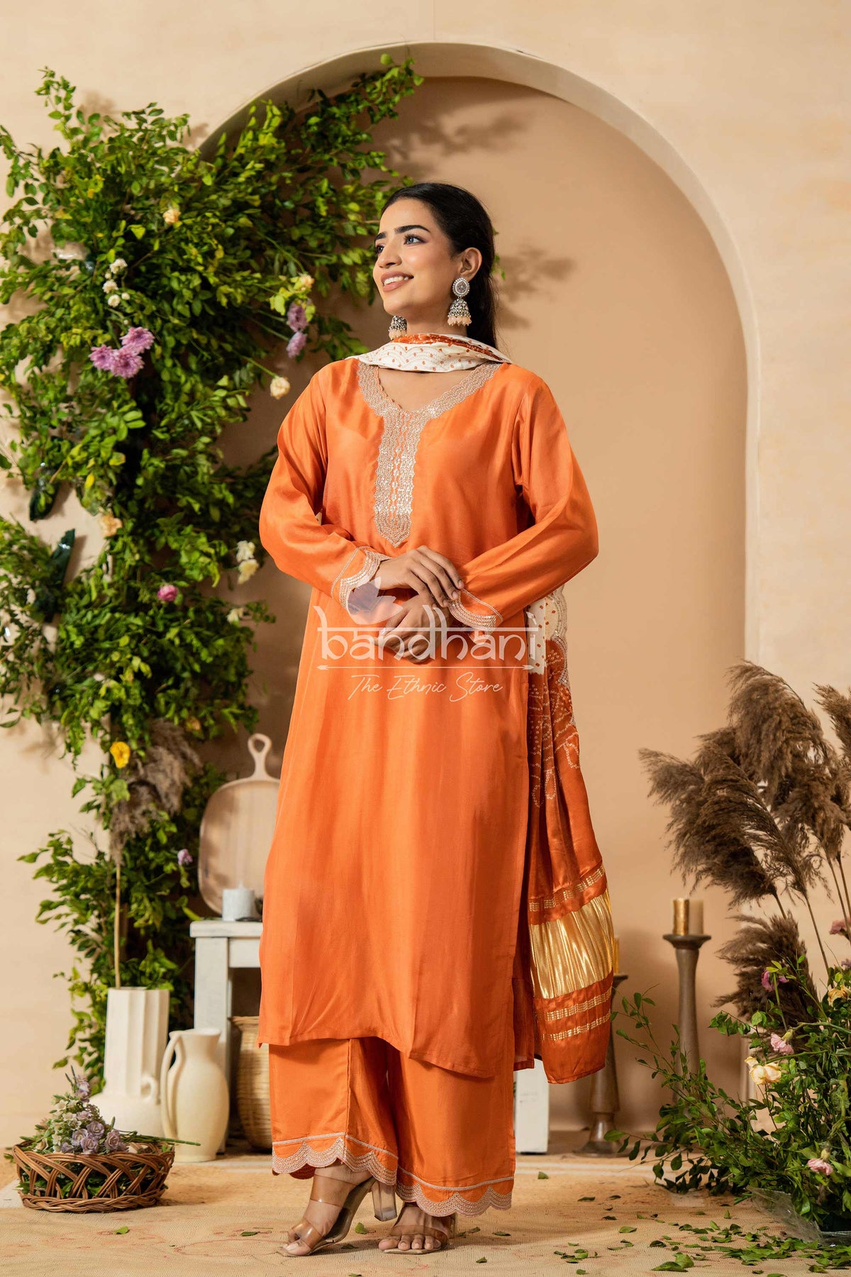 Woman in an orange traditional outfit standing in a decorated indoor setting with plants and decor.