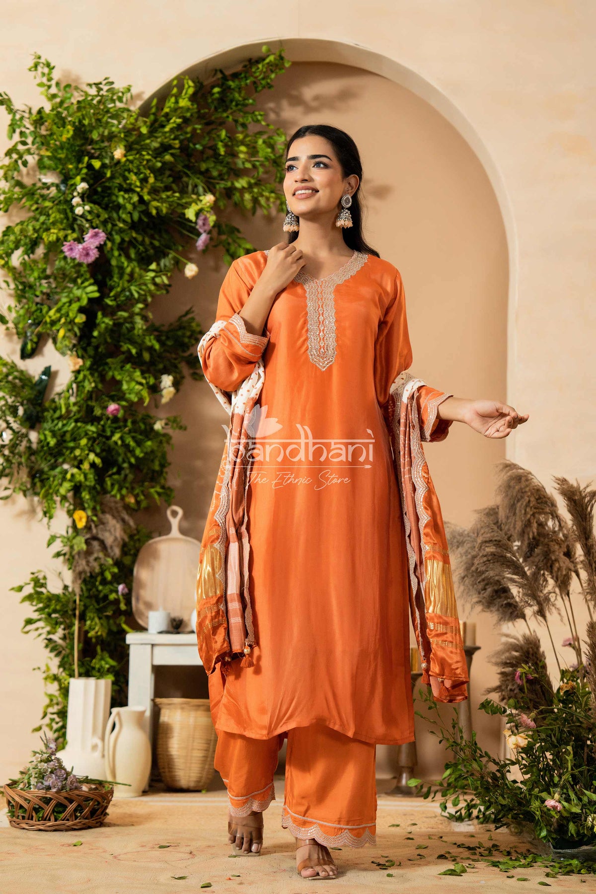 Woman in an orange traditional outfit standing in a decorative setting with plants and a beige wall.