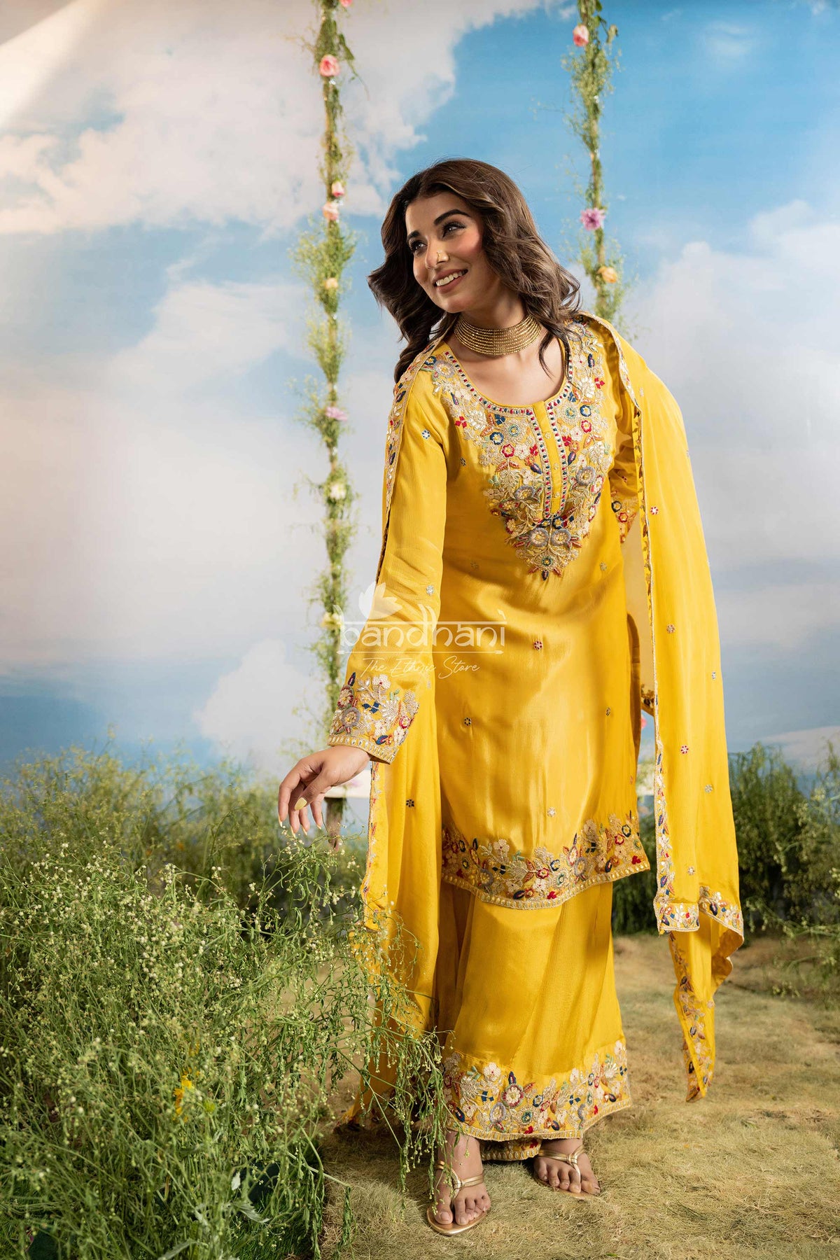 Woman in a yellow traditional outfit with floral patterns standing outdoors against a blue sky.