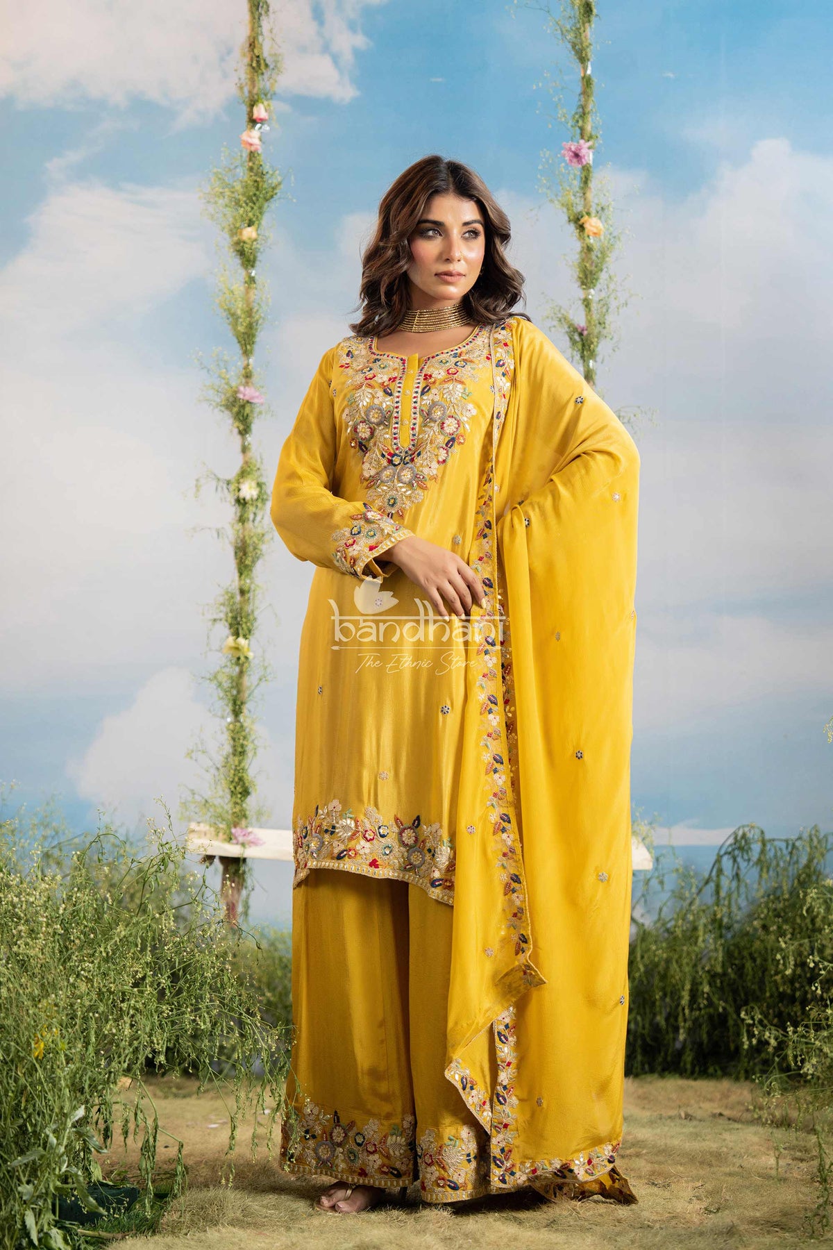Woman in a yellow traditional outfit with floral patterns standing outdoors against a blue sky.
