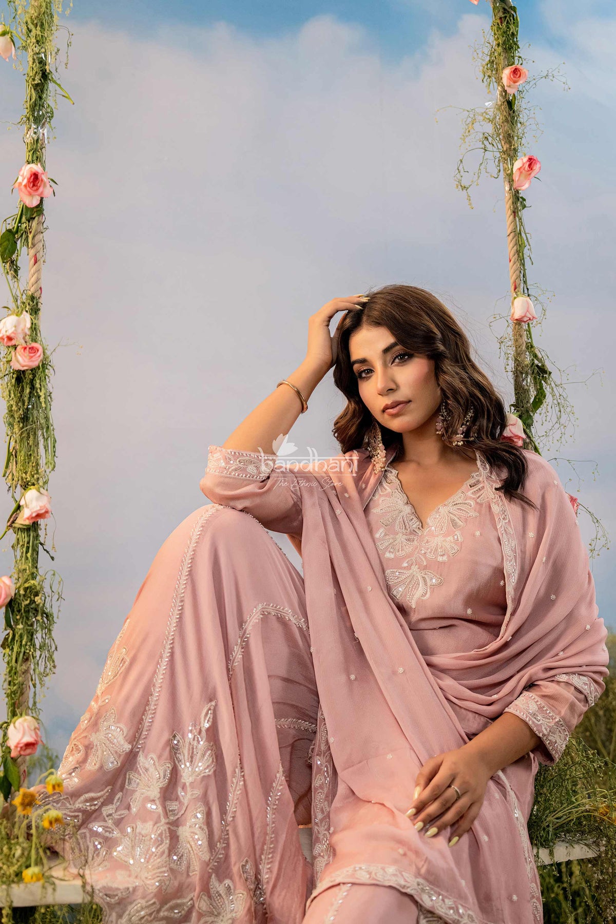 Woman in a pink traditional outfit sitting on a swing with floral decorations against a blue sky.