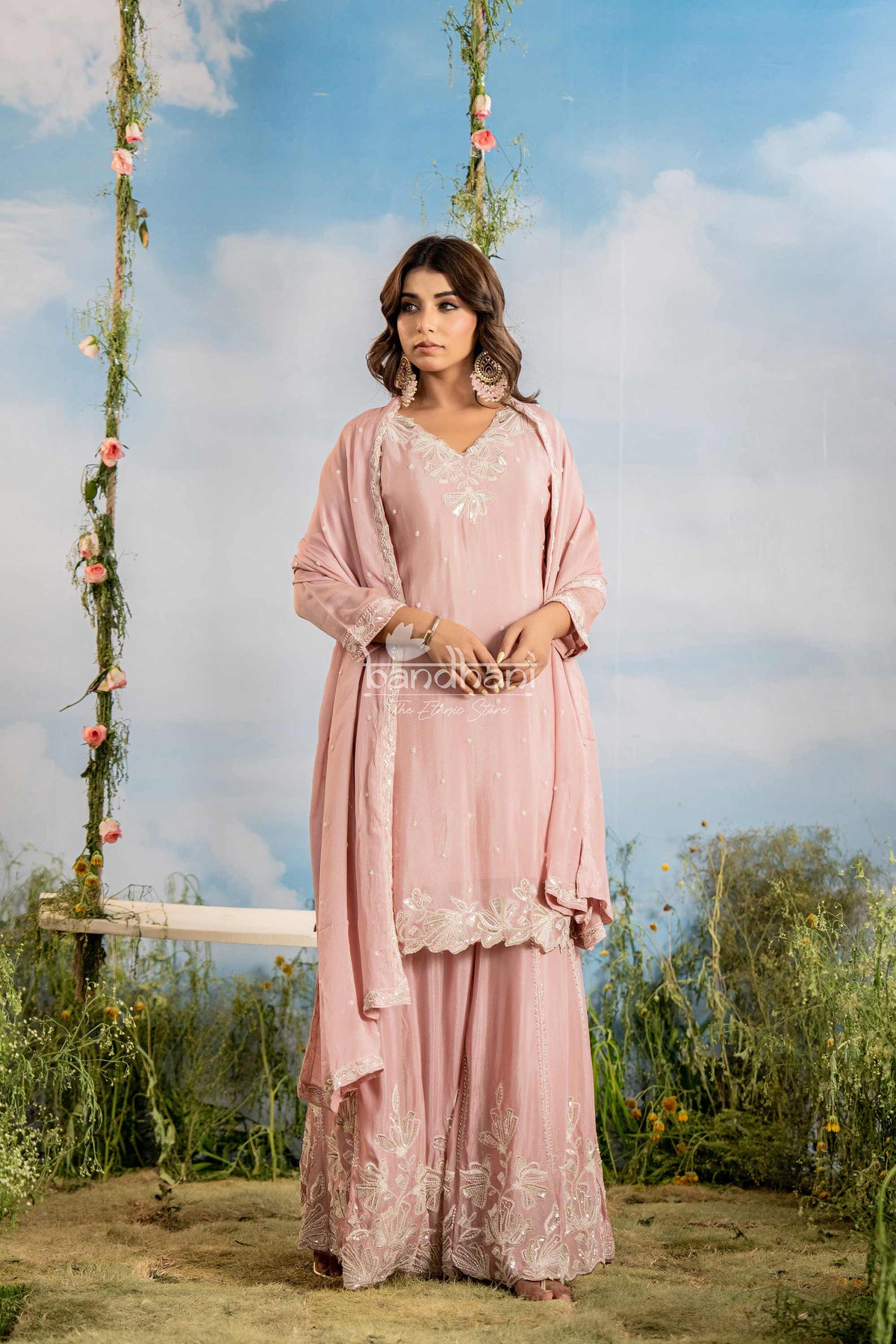 Woman in a pink traditional outfit standing on a grassy area with a swing and floral decorations in the background.
