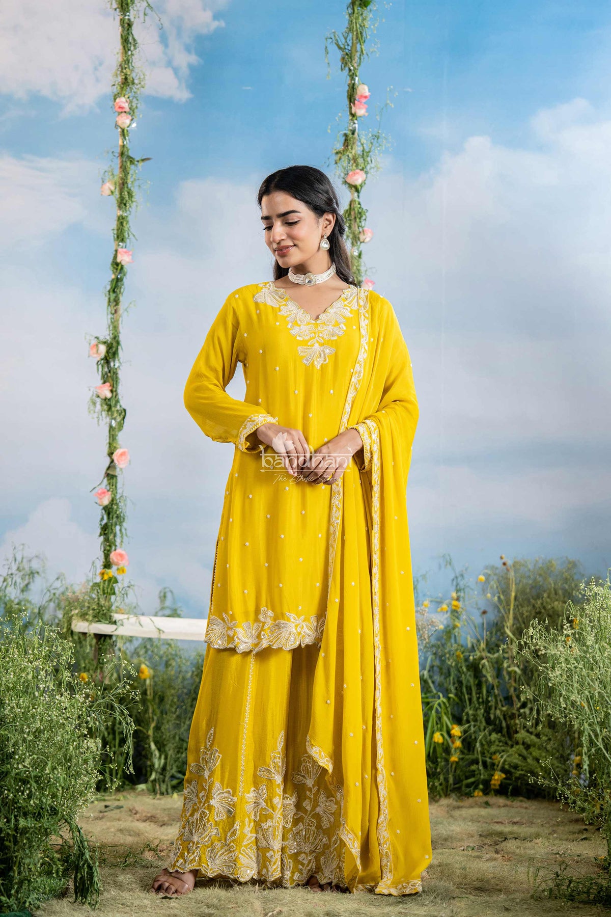 Woman in a yellow traditional outfit sitting on a swing with flowers against a blue sky.