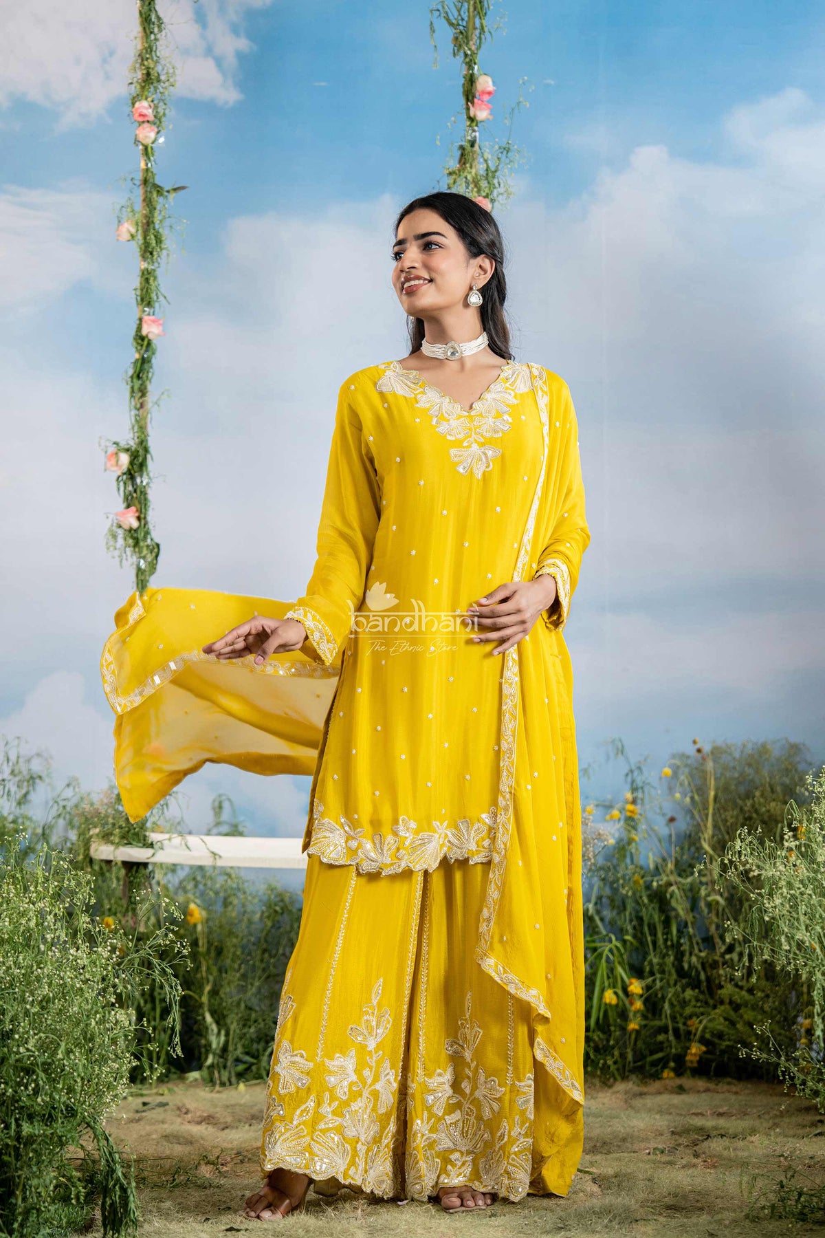 Woman in a yellow traditional outfit sitting on a swing with floral decorations against a blue sky.