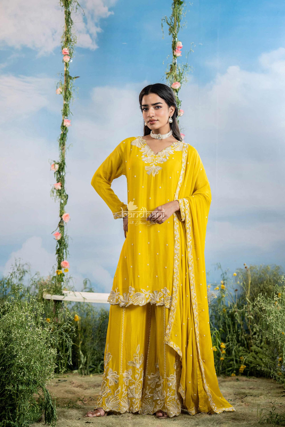 Woman in a yellow traditional outfit standing on a swing with floral decorations against a blue sky.