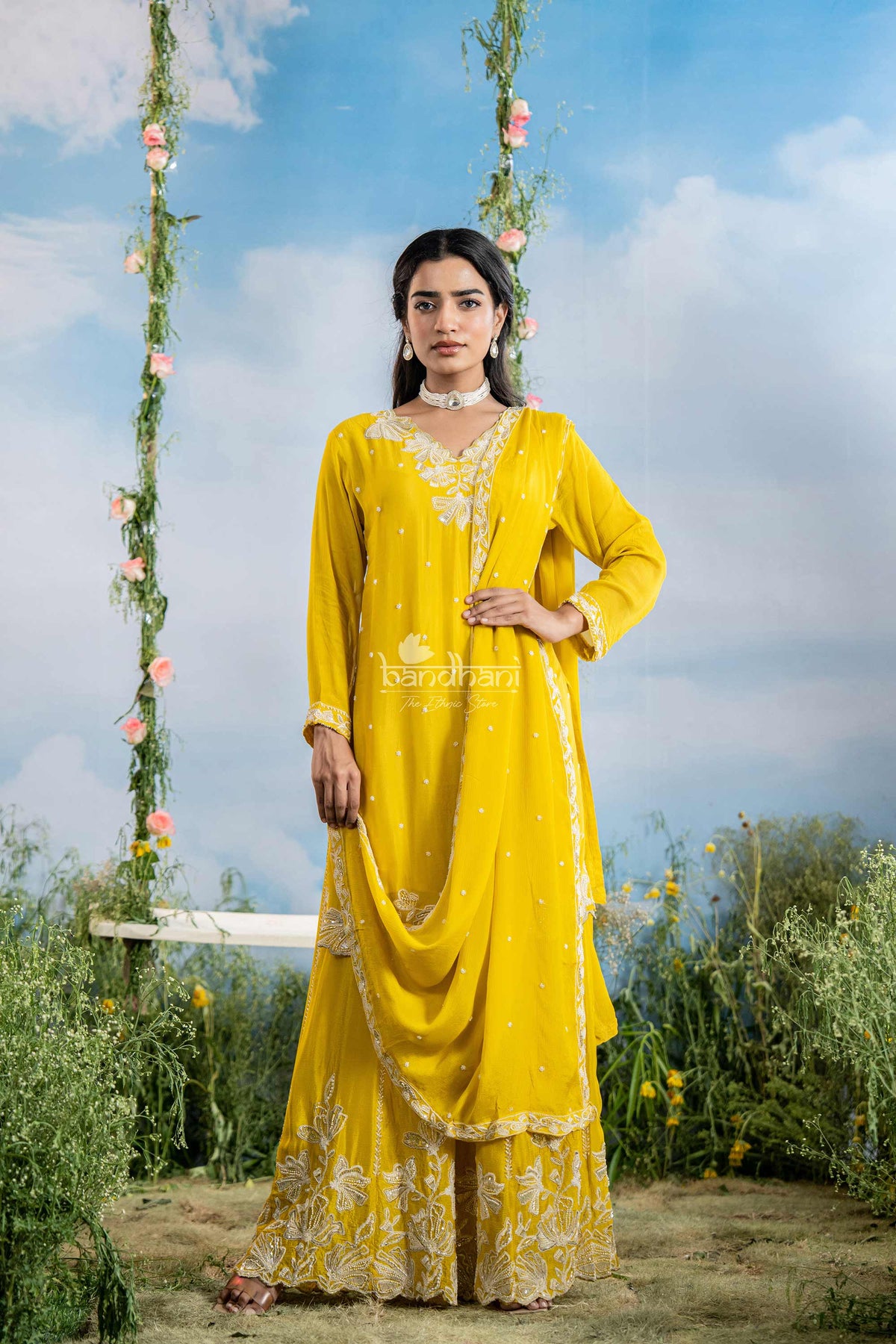 Woman in a yellow traditional outfit standing outdoors with a floral swing and blue sky in the background