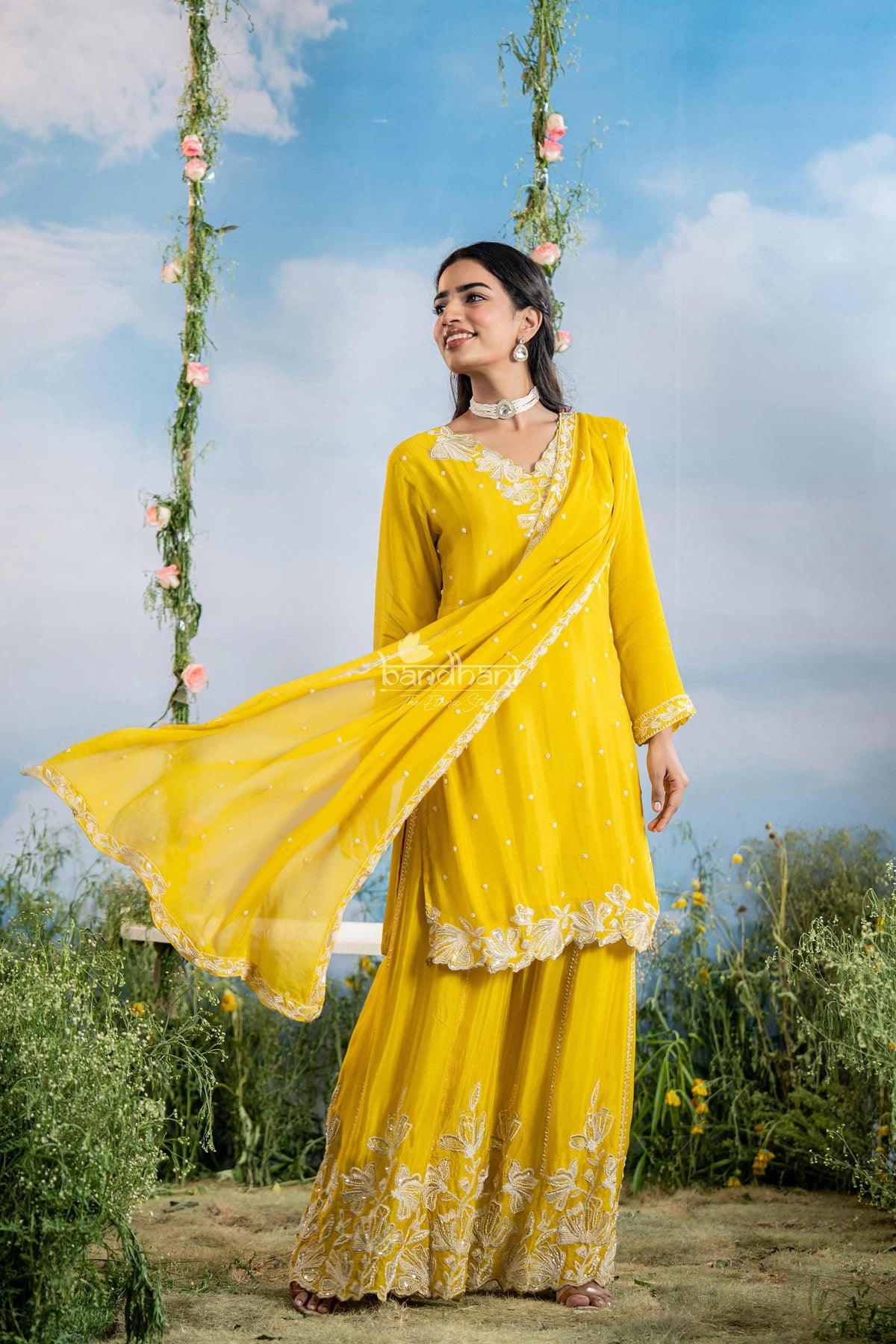 Woman in a yellow traditional outfit standing outdoors with greenery and a swing in the background