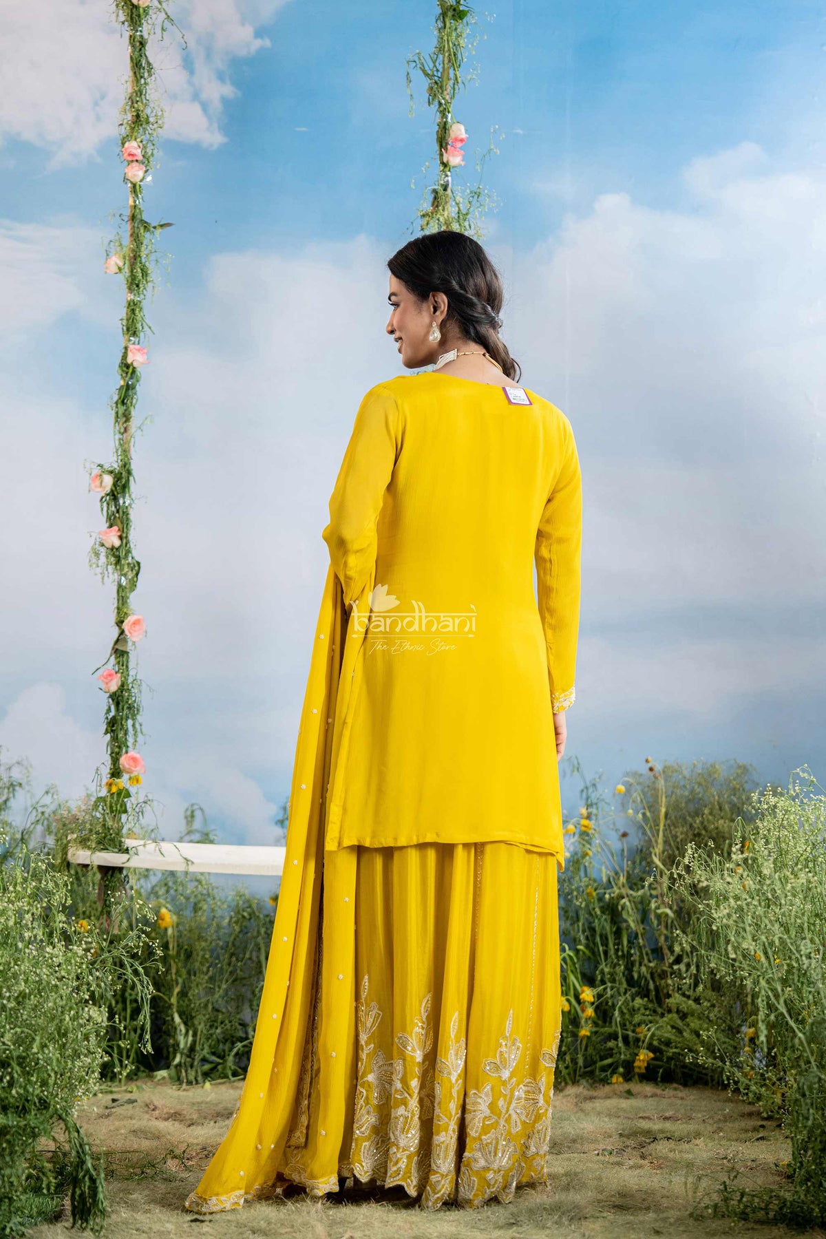 Woman in a yellow traditional outfit standing outdoors with greenery and a blue sky.