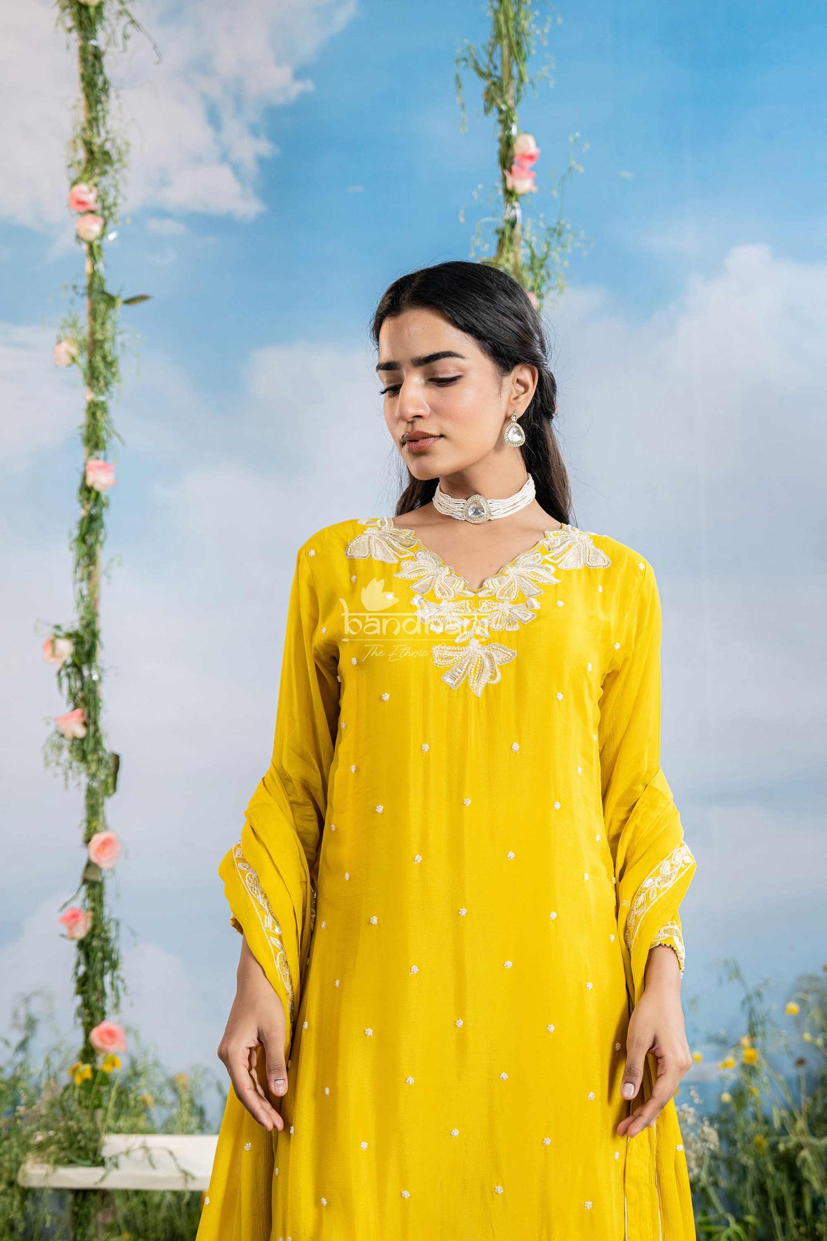 Woman wearing a yellow dress with floral patterns against a blue sky with clouds and hanging flowers.