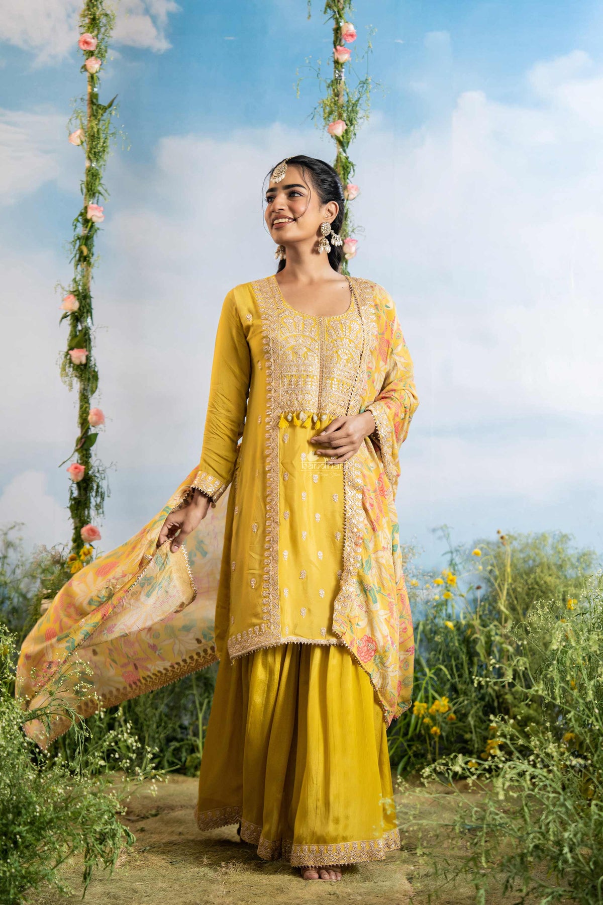 Woman in a yellow traditional outfit standing outdoors with flowers and greenery around.