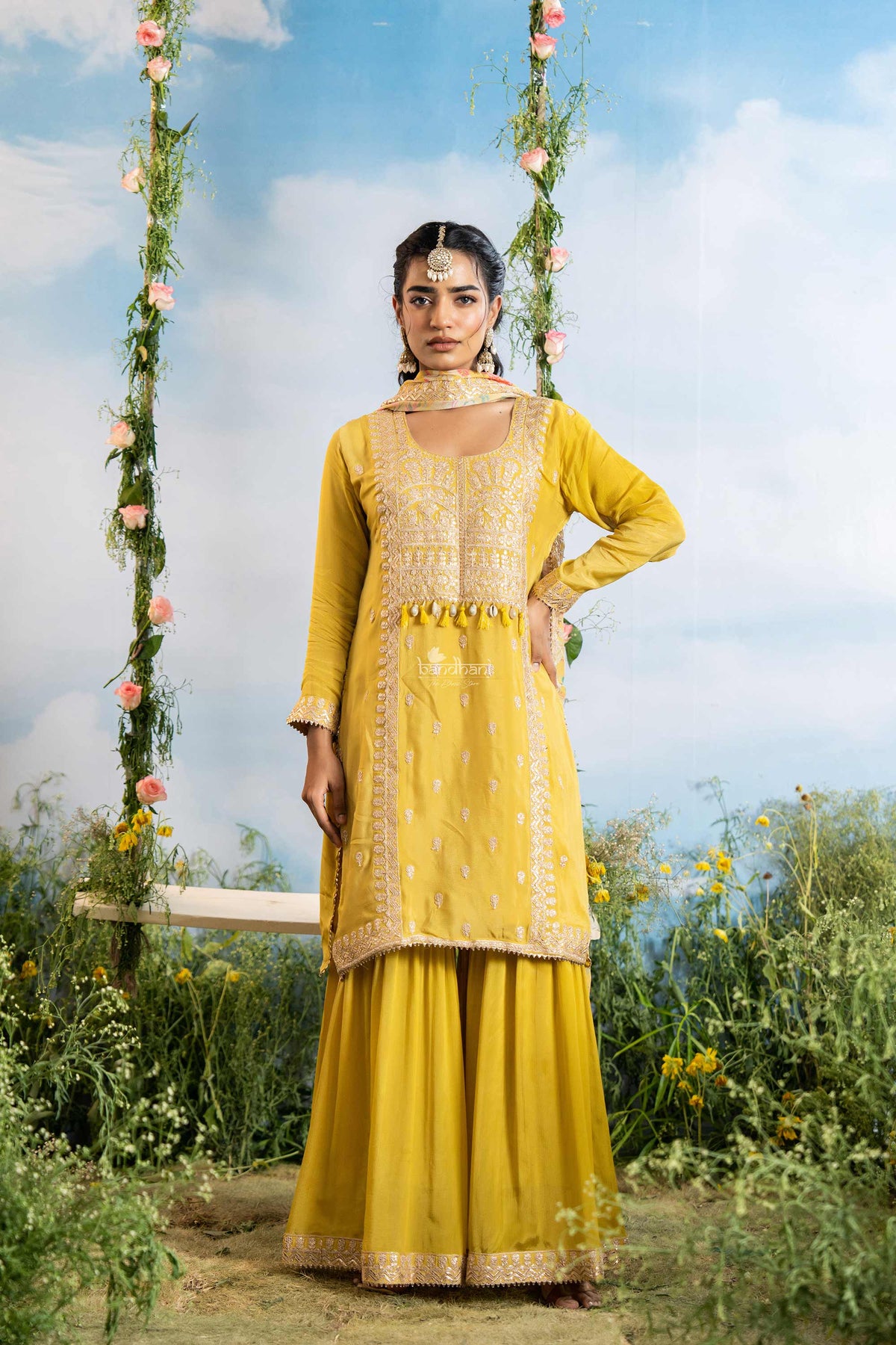 Woman in a yellow traditional outfit standing outdoors with a swing and flowers in the background