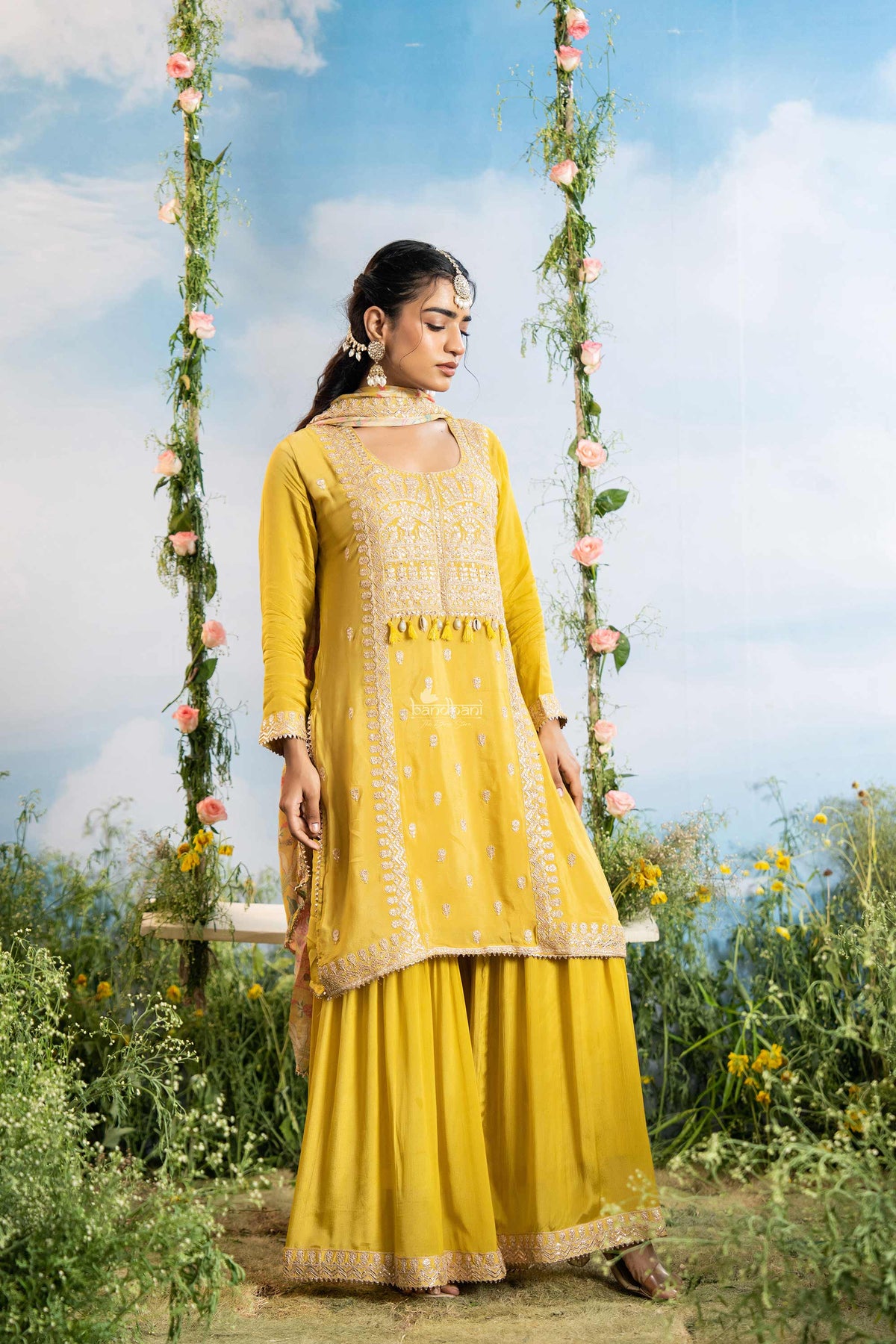 Woman in a yellow traditional outfit standing on a swing with floral decorations against a blue sky.