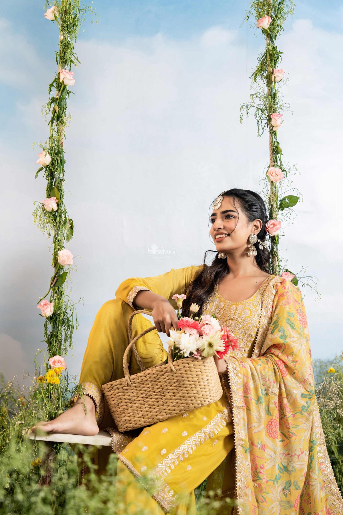 Woman in a yellow saree with floral decorations on a swing against a blue sky.