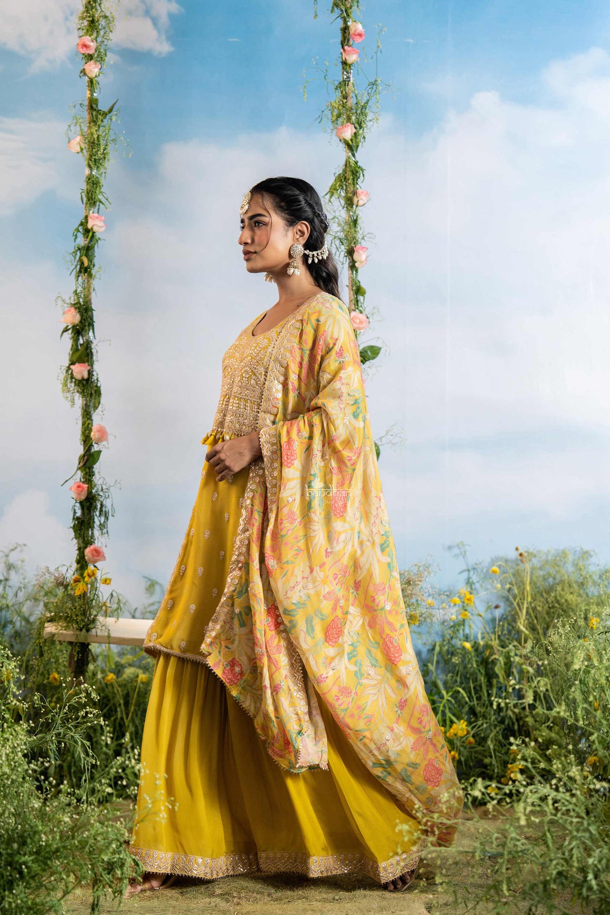 Woman in a yellow and floral traditional outfit standing outdoors with greenery and flowers around.