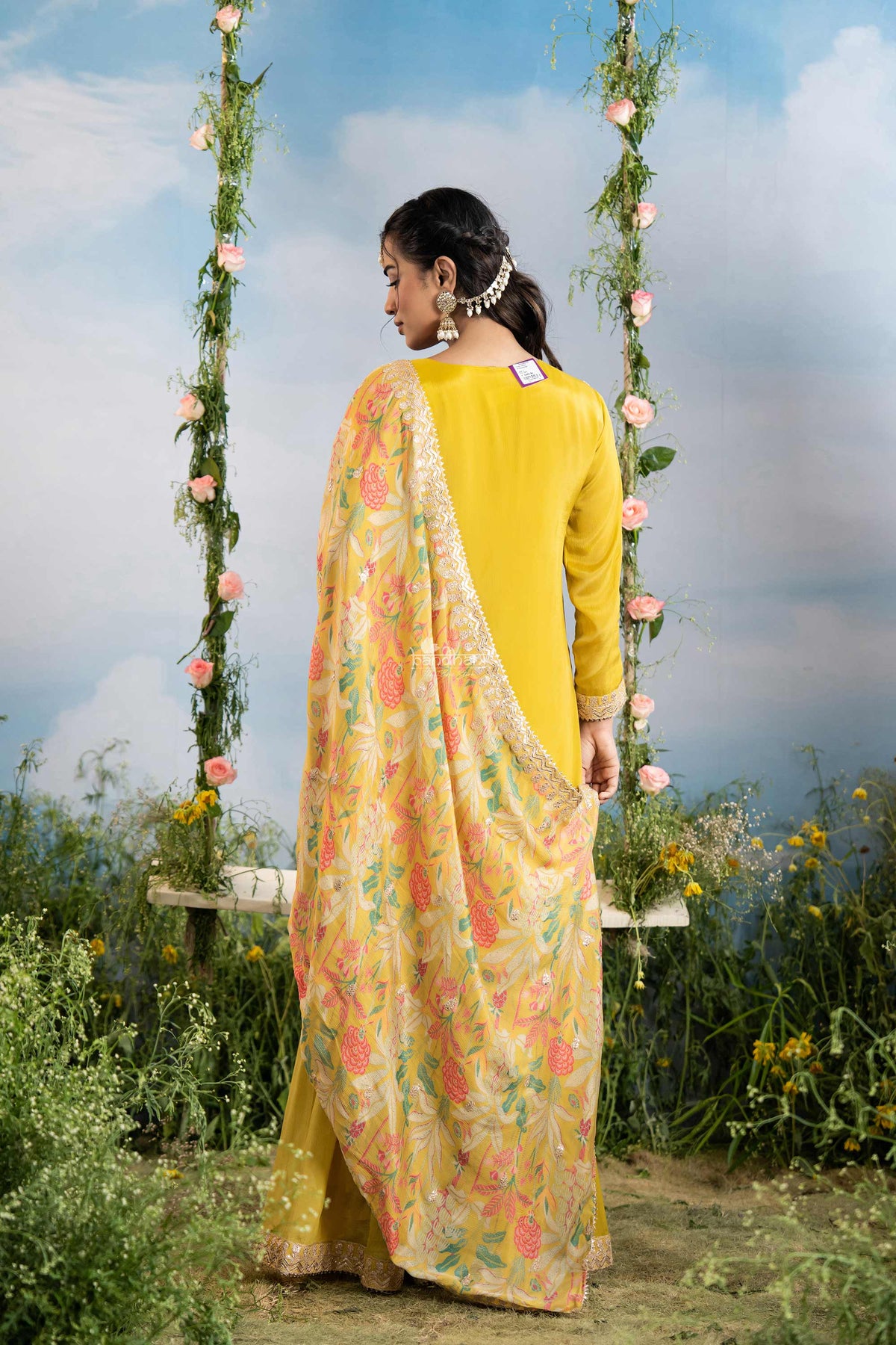 Woman in a yellow outfit with a floral dupatta sitting on a swing in a natural setting.