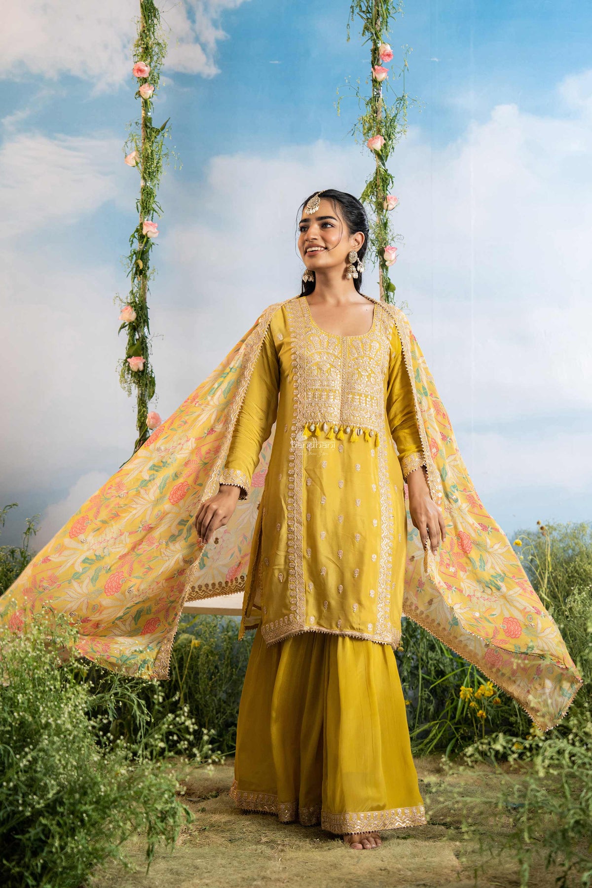 Woman in a yellow traditional outfit with a floral dupatta against a blue sky with clouds and greenery.