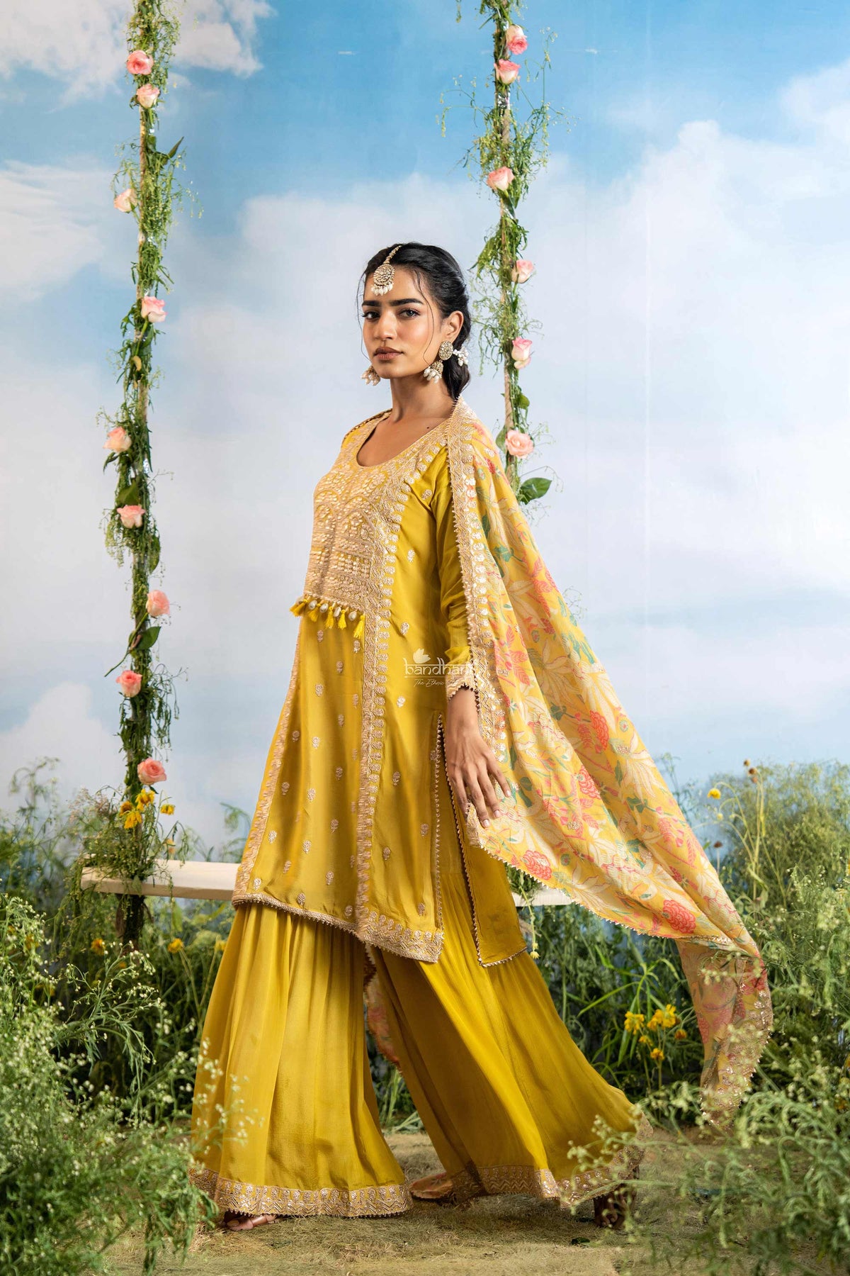 Woman in a yellow traditional outfit standing on a swing with floral decorations against a blue sky.