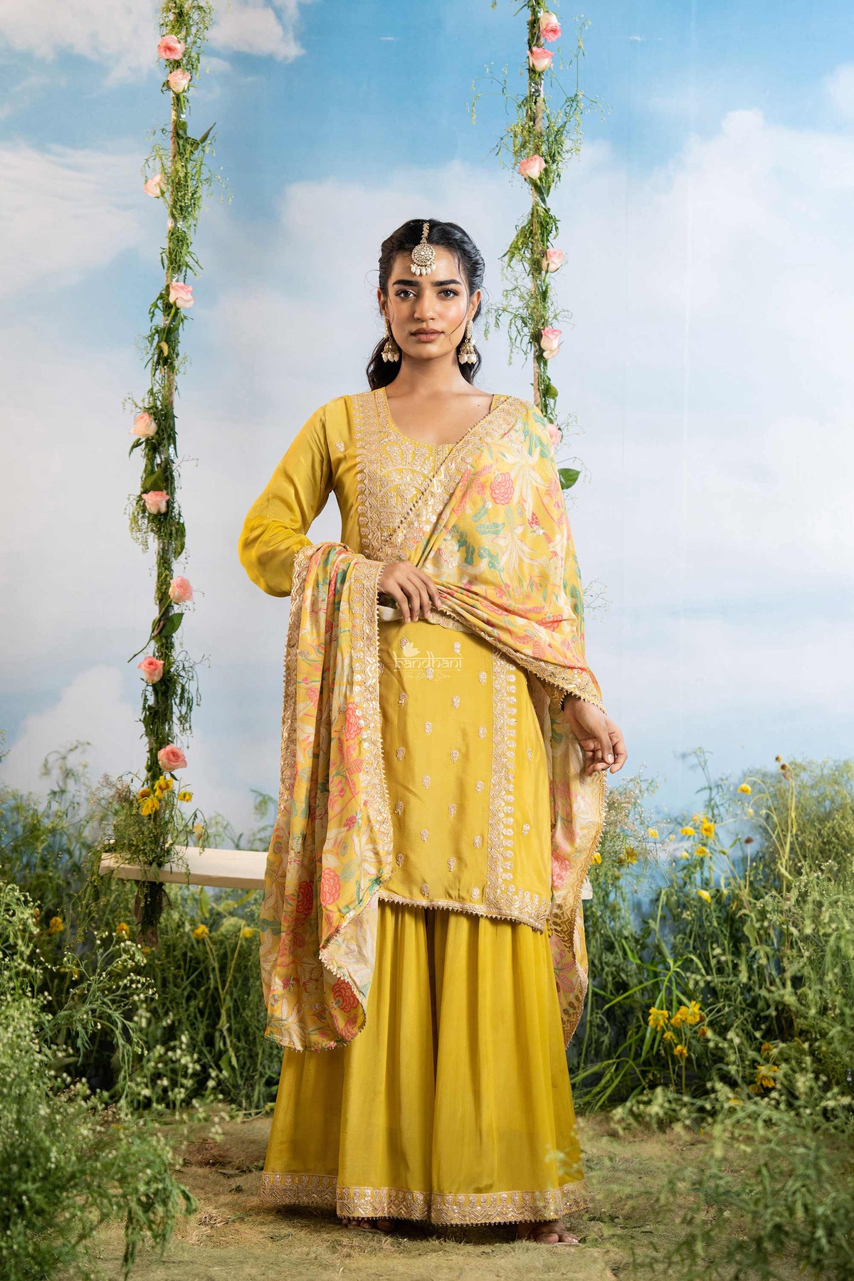 Woman in a yellow traditional outfit standing on a swing with floral decorations against a blue sky.