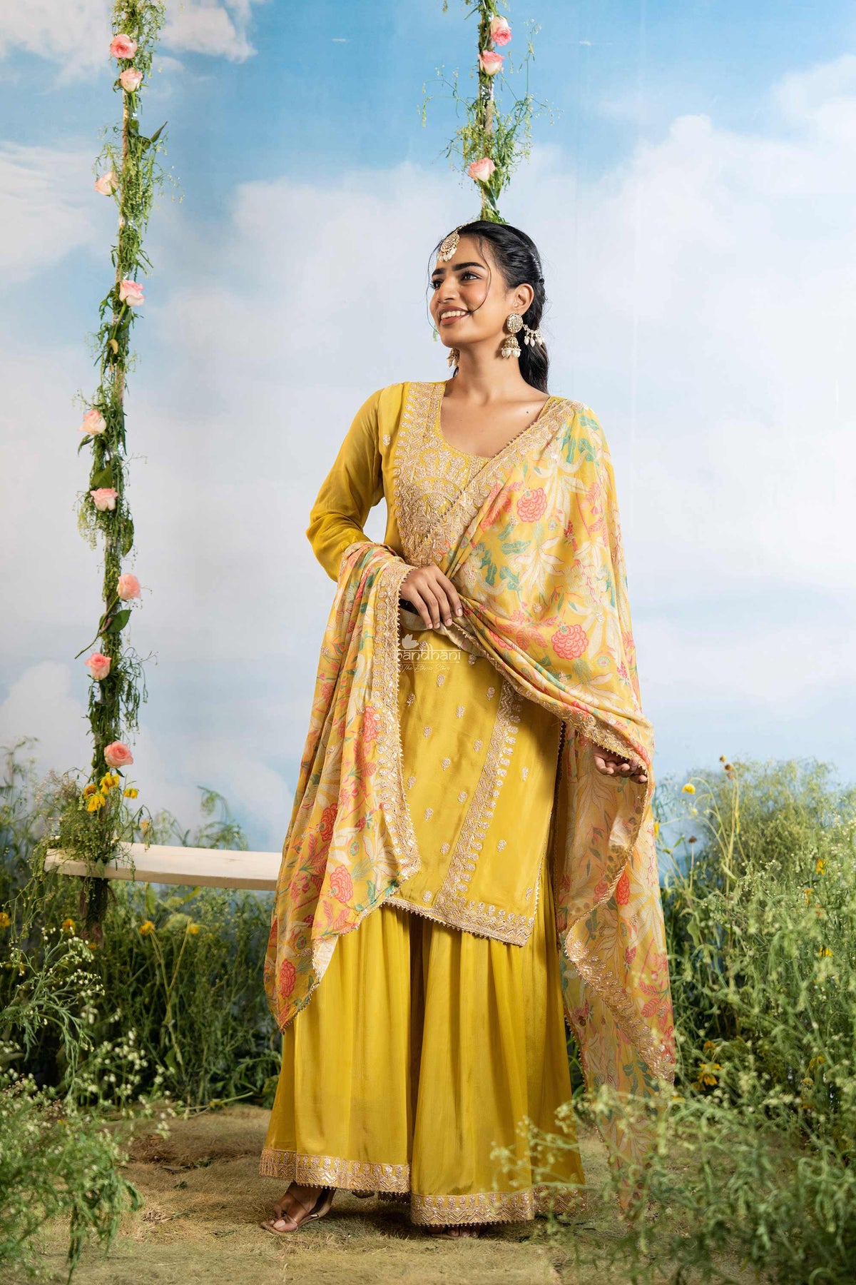 Woman in a yellow traditional outfit with a floral dupatta standing outdoors with a scenic background.