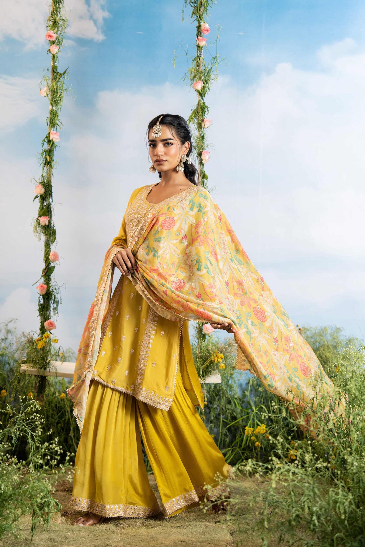 Woman in a yellow traditional outfit with a floral dupatta standing outdoors with a blue sky and greenery in the background.