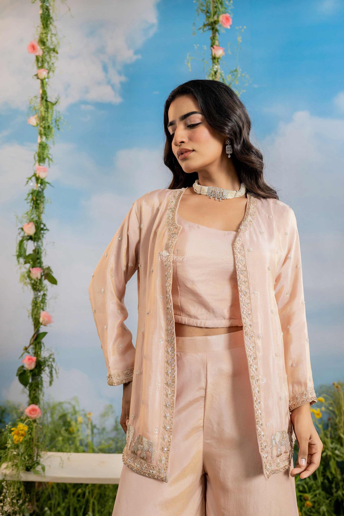 Woman in a peach traditional outfit standing against a scenic background with flowers and sky.