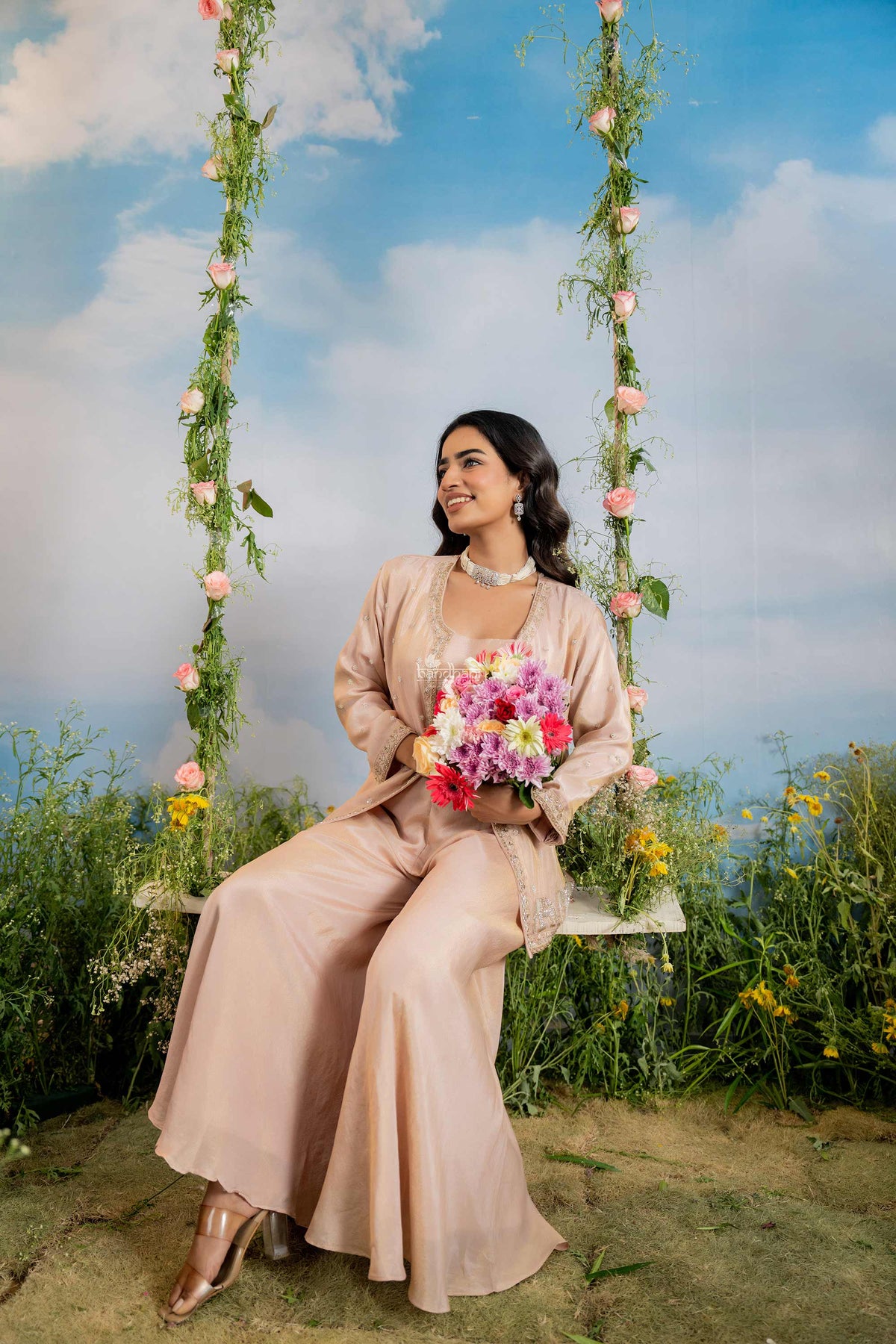 Woman in a peach outfit sitting on a floral swing with a colorful bouquet against a blue sky.