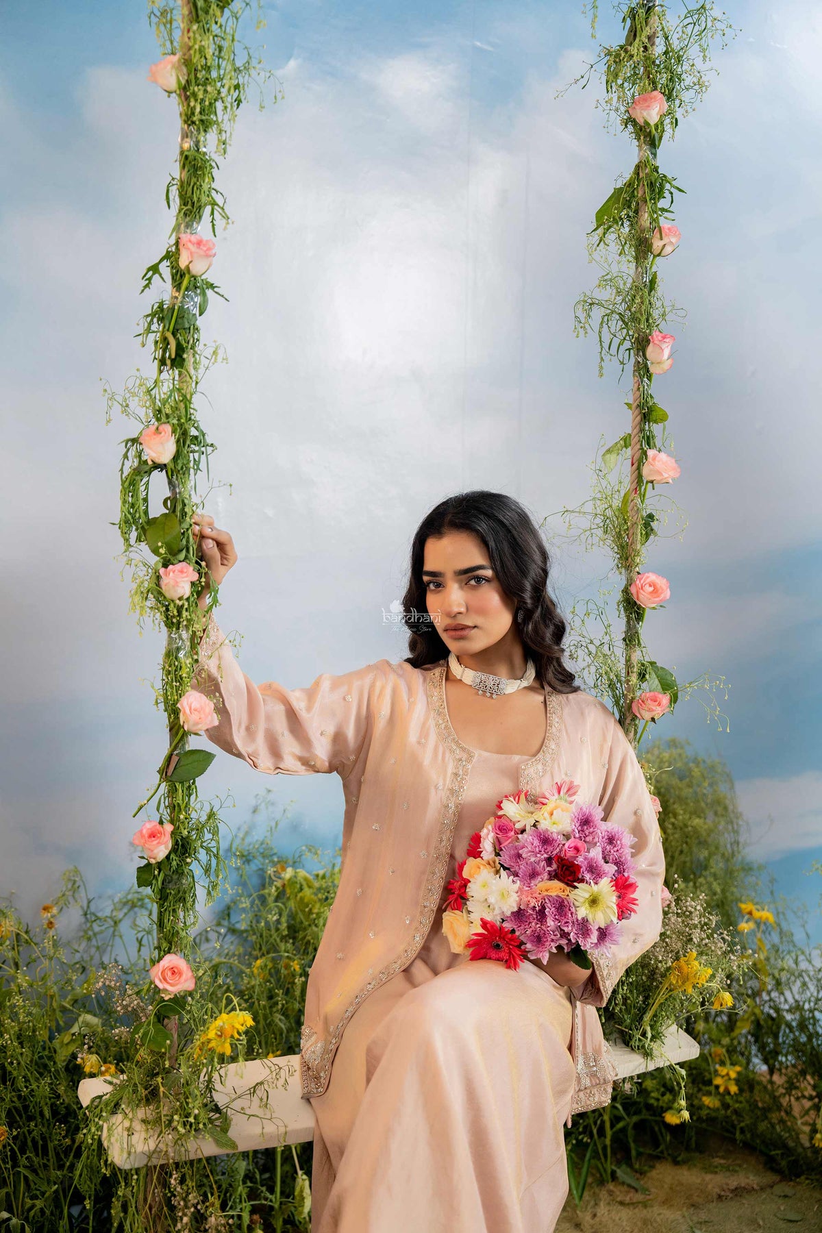 Woman in a light pink traditional outfit holding flowers, sitting on a floral swing with a scenic background.