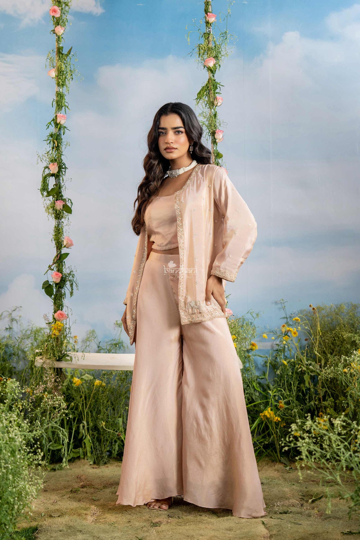 Woman in a beige outfit sitting on a swing in a garden with a blue sky.