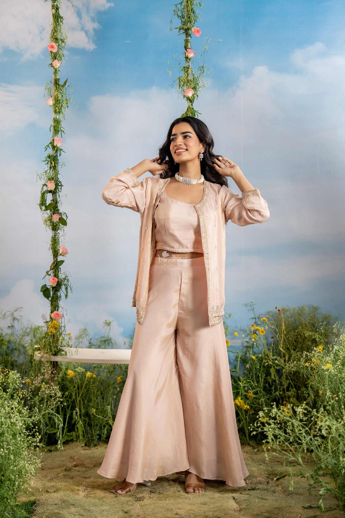 Woman in a beige outfit standing in a garden with flowers and a blue sky.