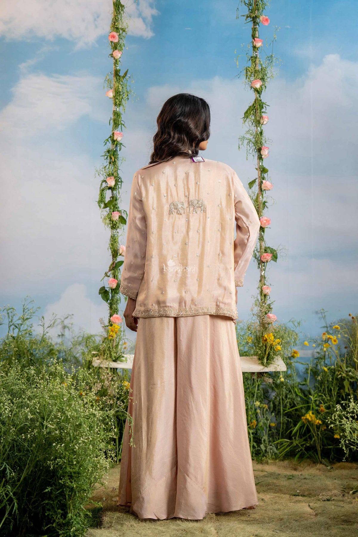 Person wearing a light pink traditional outfit standing in a garden with flowers and sky in the background
