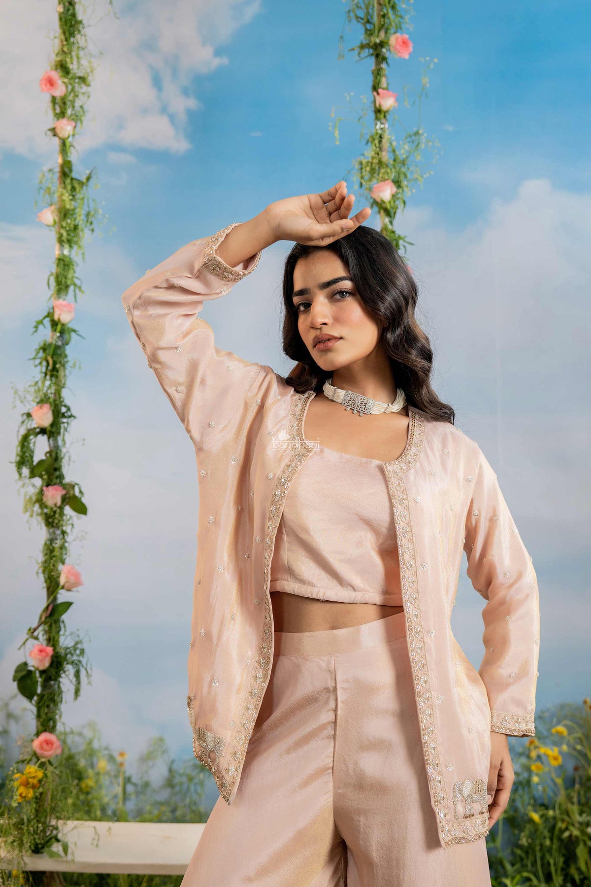 Woman in a light pink traditional outfit standing outdoors with floral decorations and a blue sky.