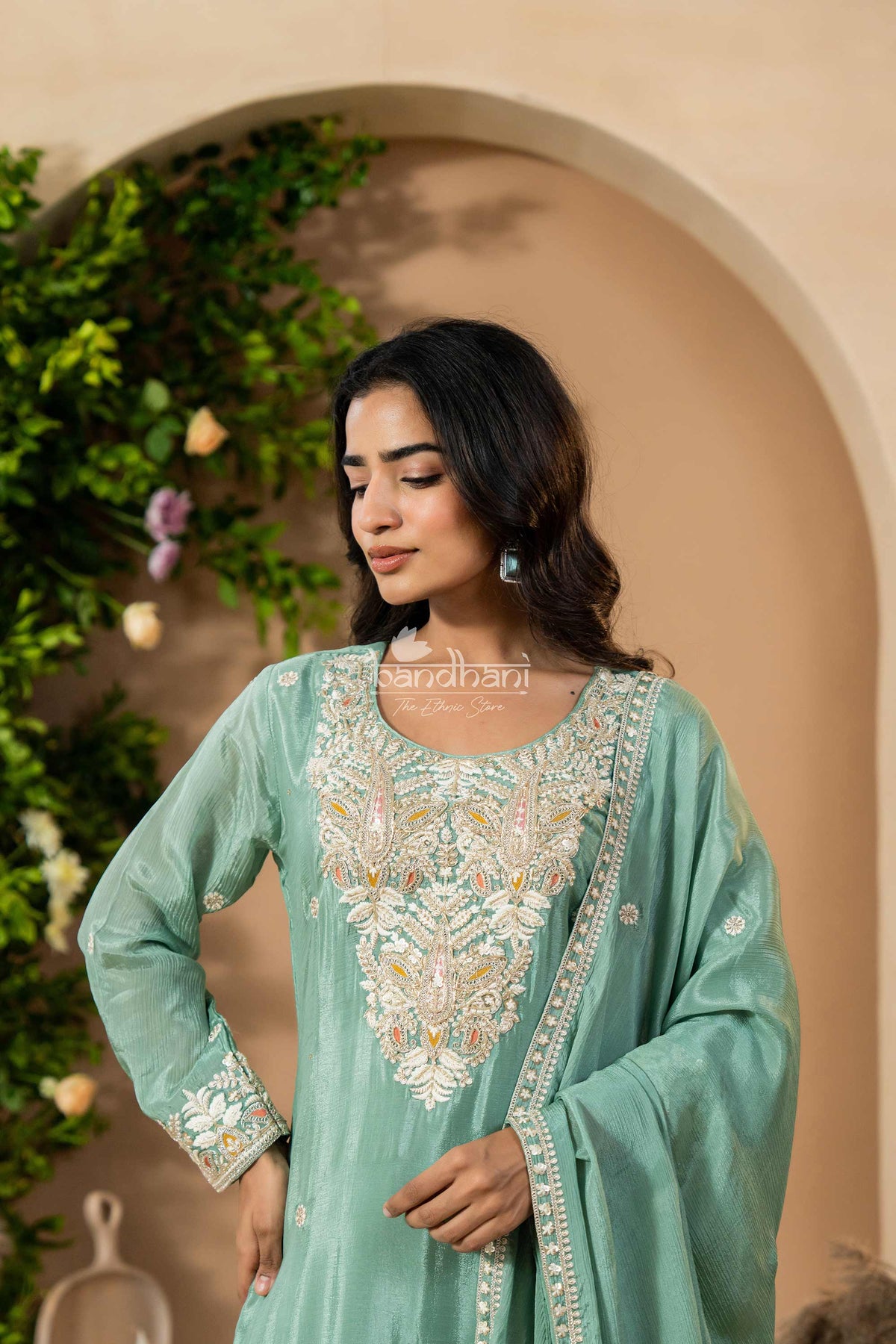 Woman wearing a light green embroidered traditional outfit with a decorative arch and plants in the background