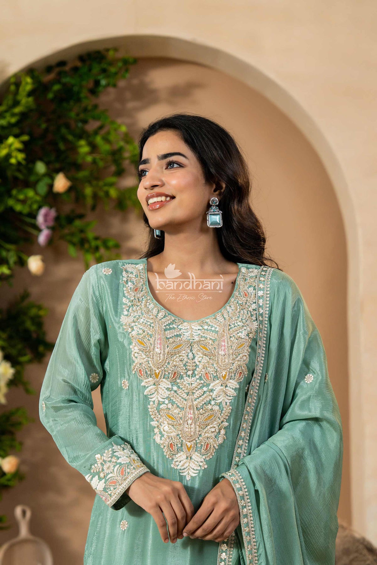 Woman wearing a light blue embroidered traditional outfit with a beige wall and greenery in the background