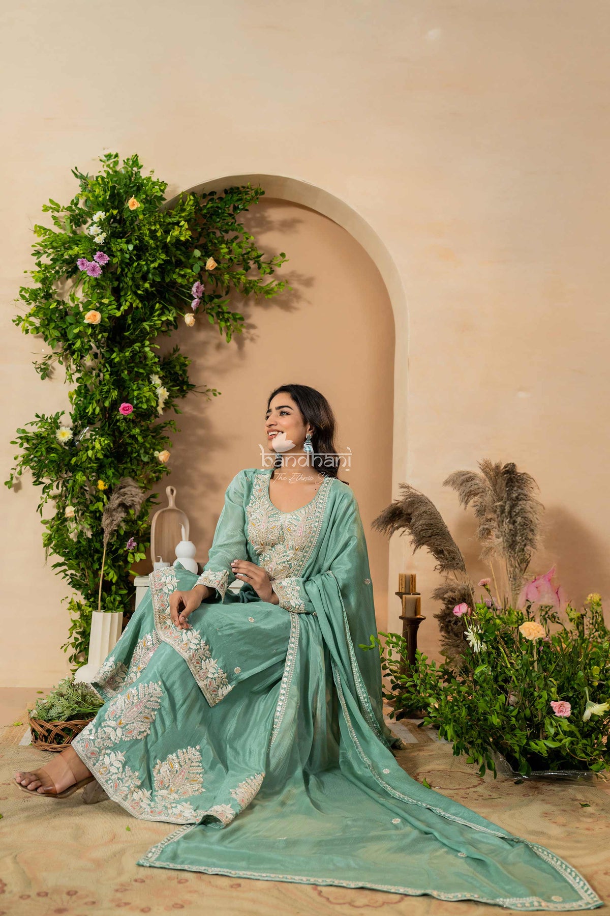 Woman in a light blue traditional outfit sitting in a decorated indoor setting with plants and flowers.