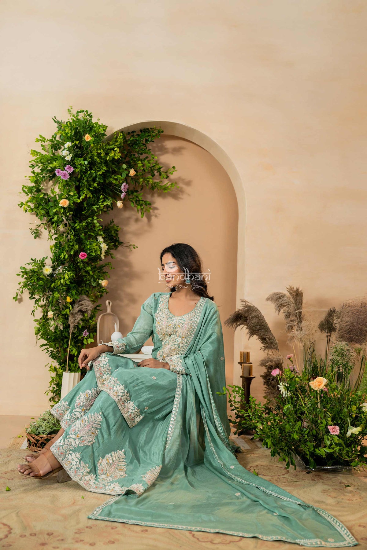 Woman in a light blue traditional outfit sitting in a decorated outdoor setting with plants and flowers.