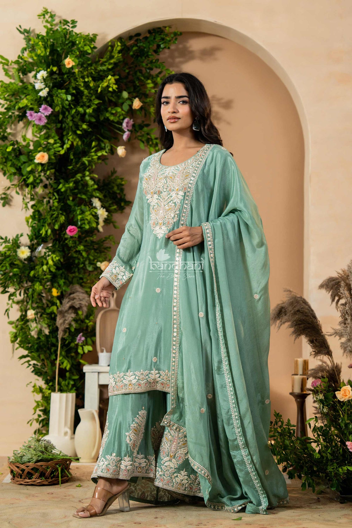 Woman in a light green traditional outfit with floral patterns standing in a decorated indoor setting.