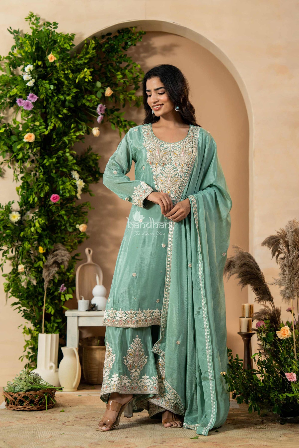 Woman in a light blue traditional outfit standing in a decorated indoor setting with plants and flowers.