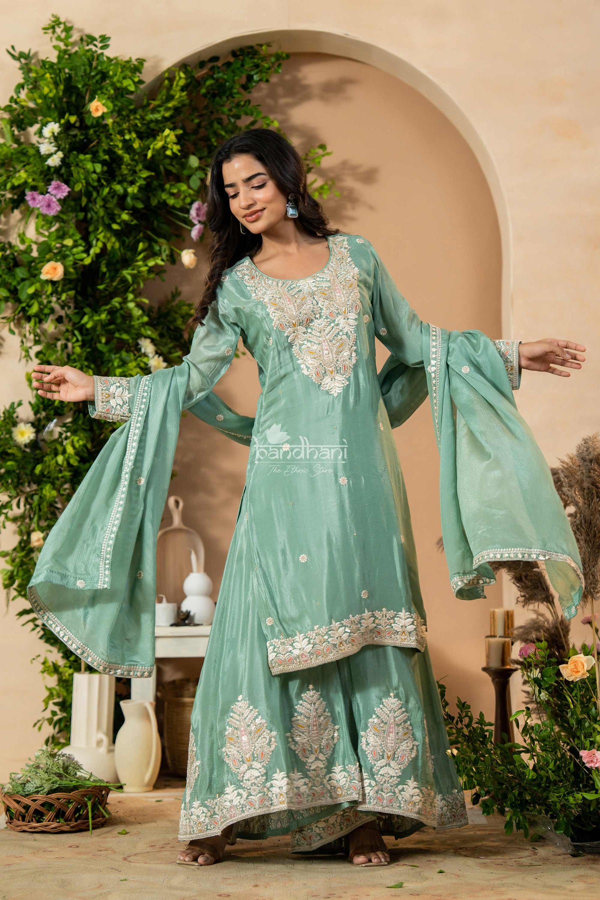 Woman in a light green embroidered traditional outfit standing in a decorated indoor setting with plants.