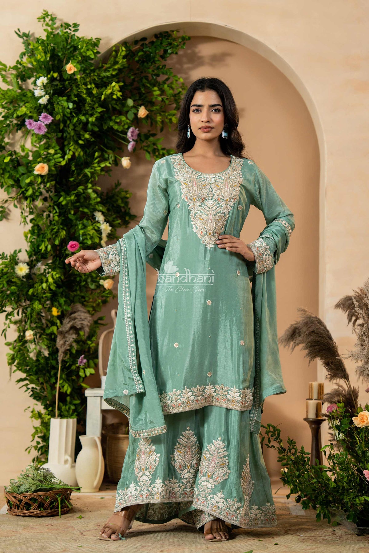 Woman in a light green embroidered traditional outfit standing in front of decorative plants and archway.