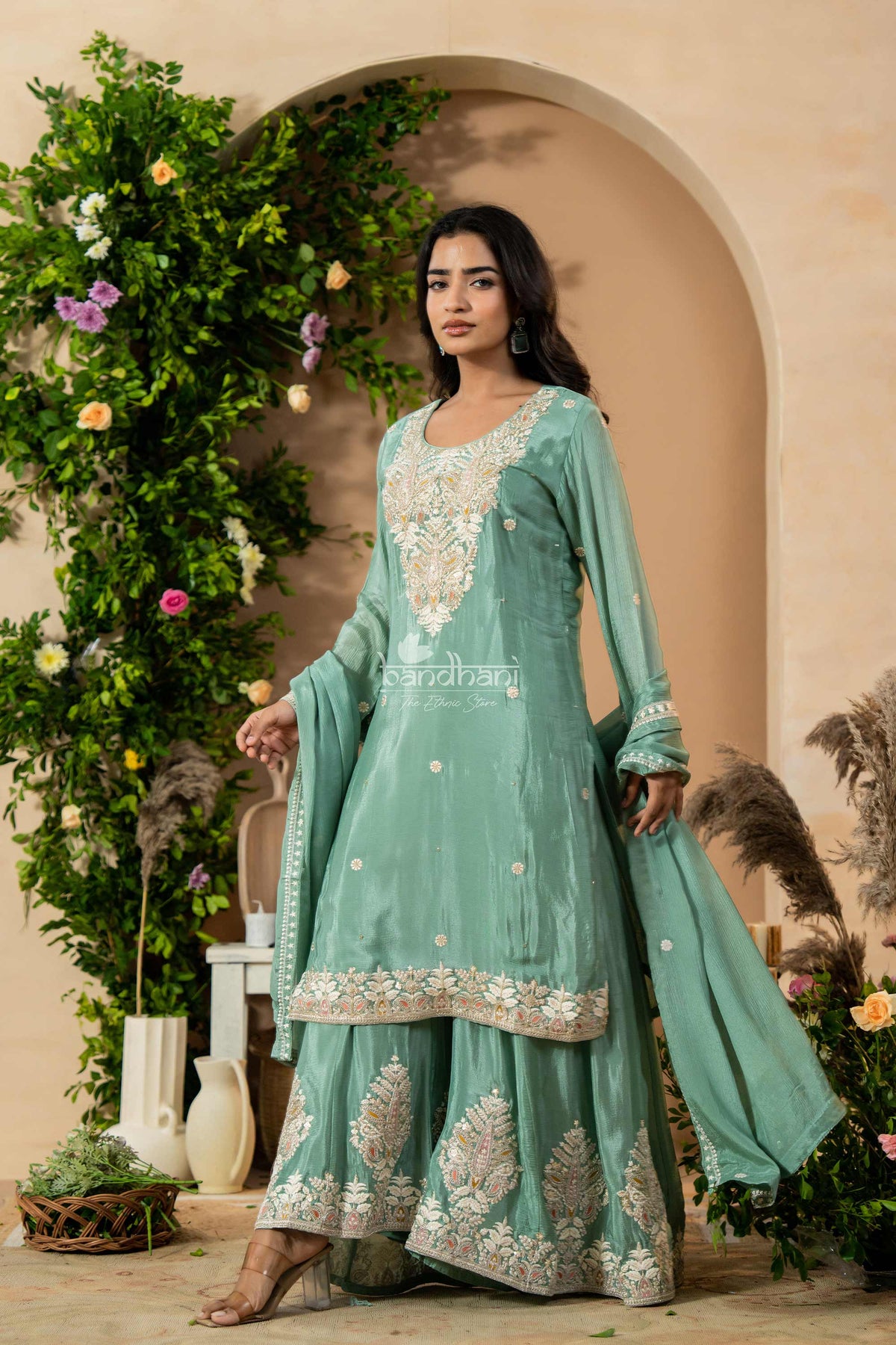 Woman in a teal embroidered outfit standing in front of decorative plants and archway.