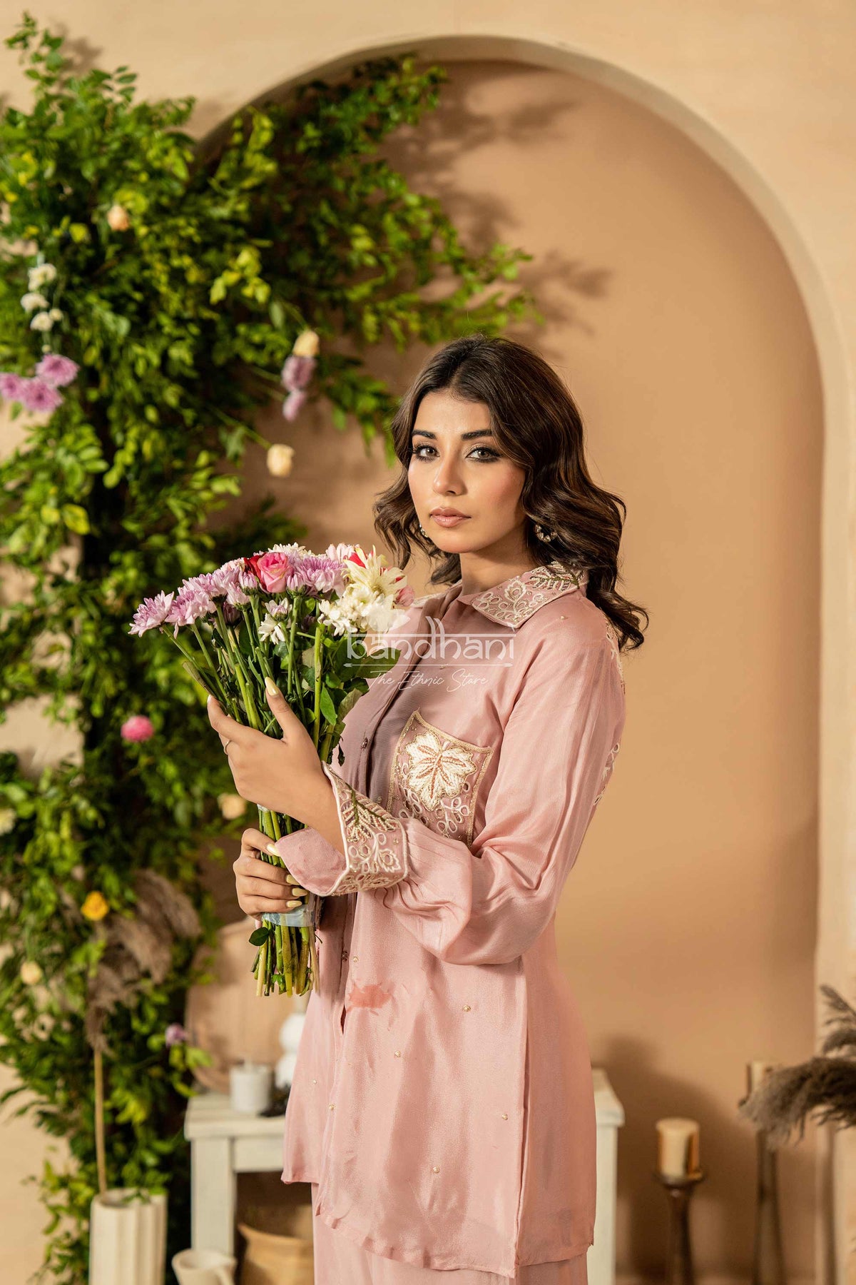 Woman in a pink dress holding flowers in an indoor setting with decorative plants.