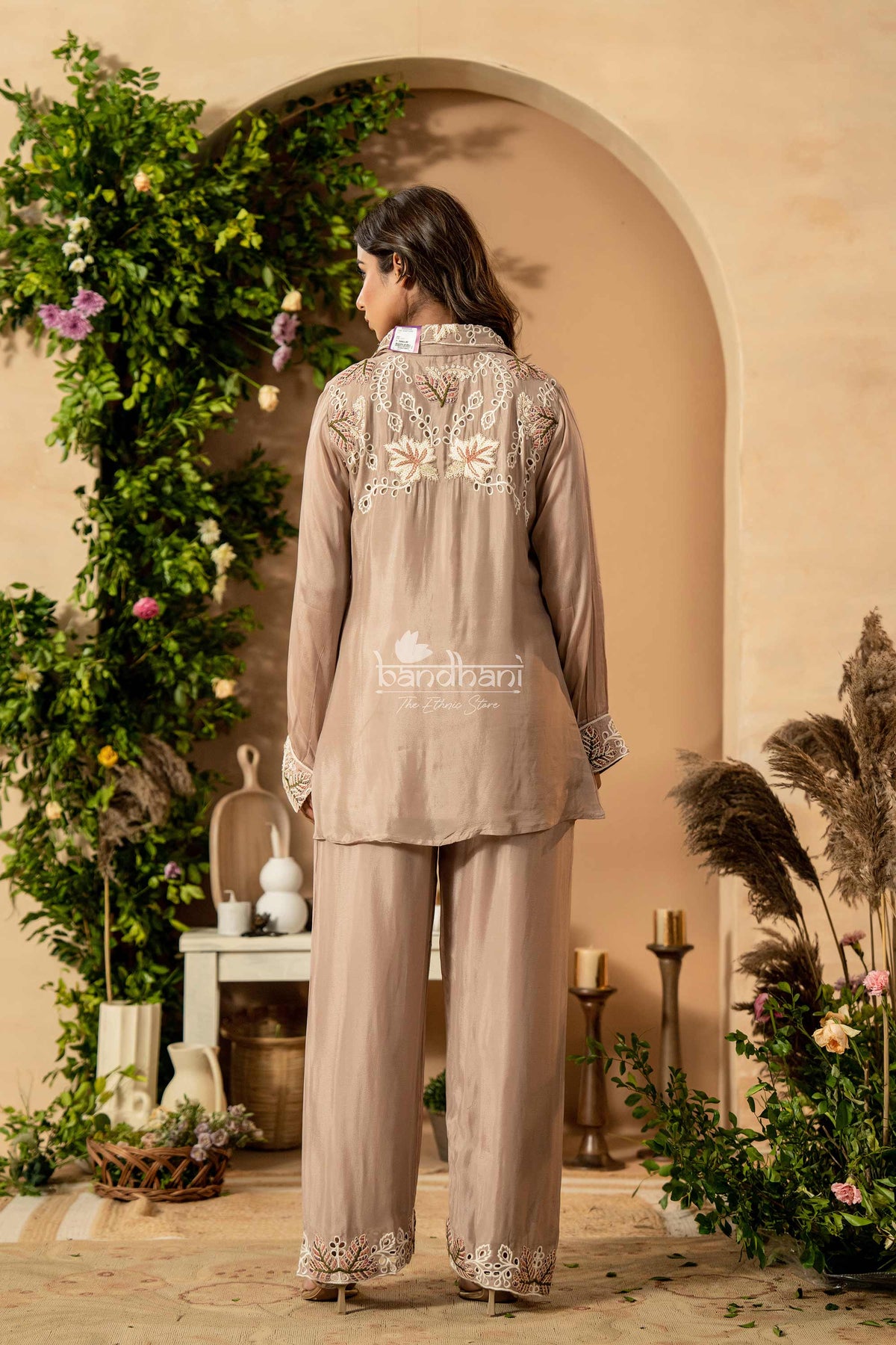 Woman wearing a beige embroidered outfit standing in a decorative indoor setting with plants and candles.