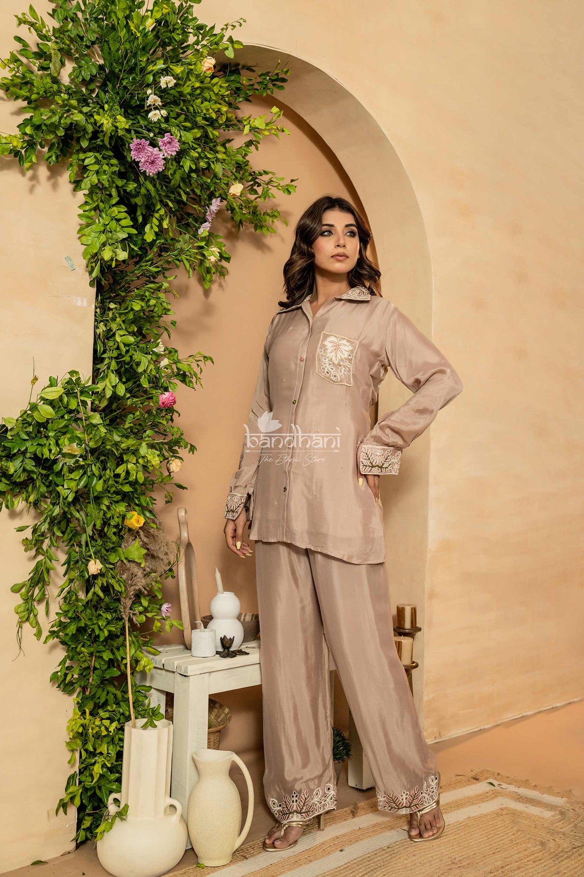Woman in a beige outfit standing next to a decorative arch with plants and flowers.