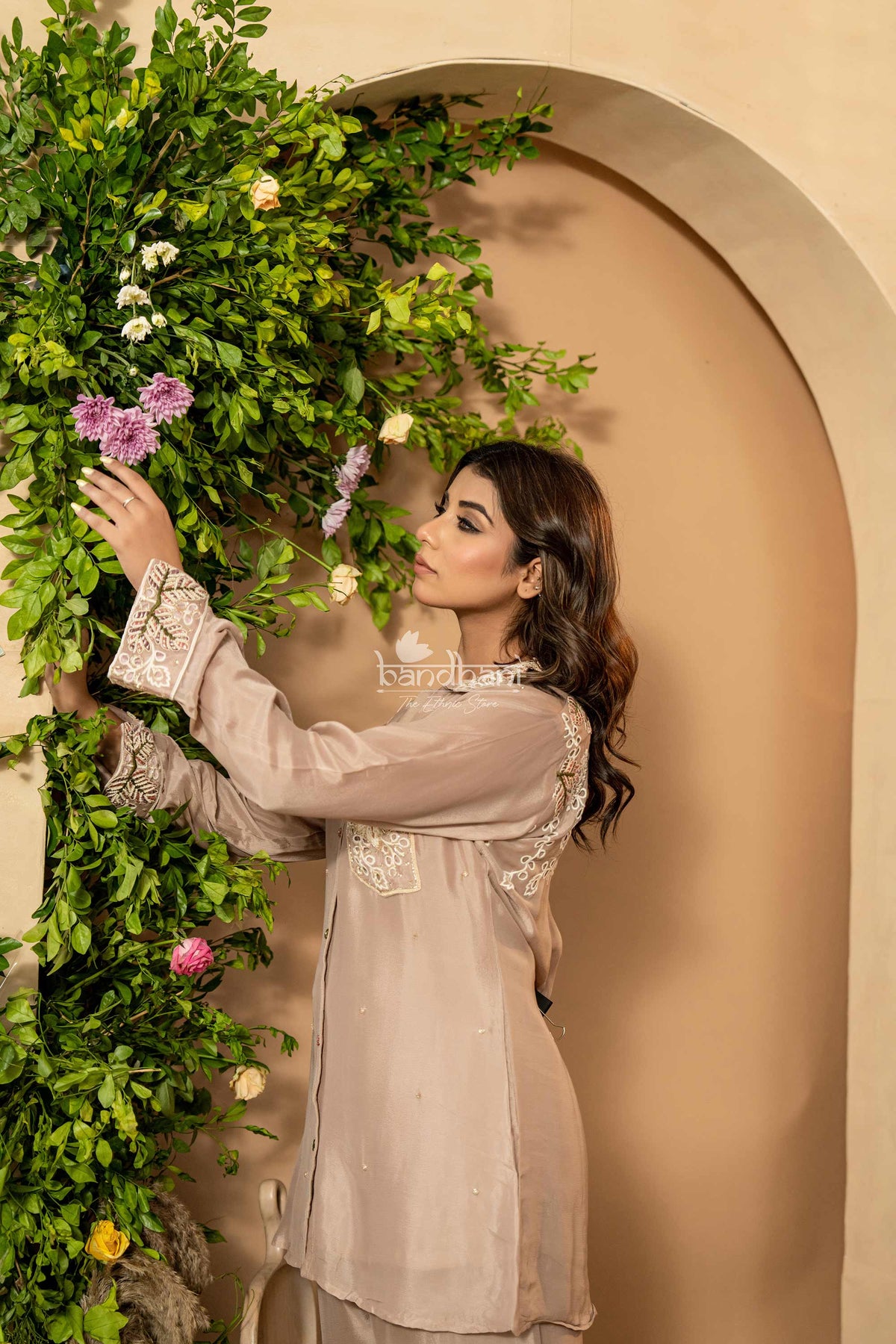 Woman in a beige dress standing next to a wall with green foliage and flowers.