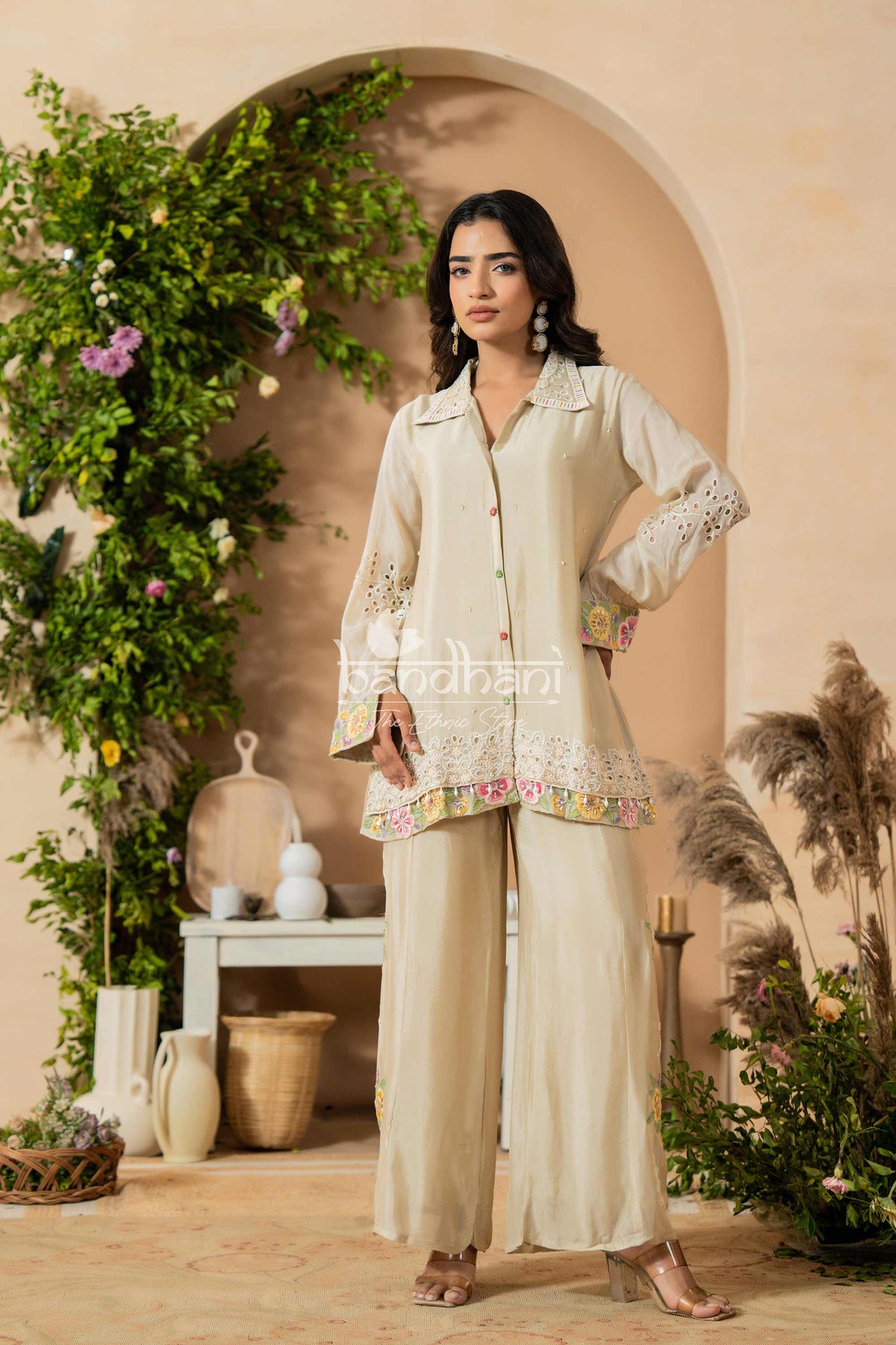 Woman in a cream traditional outfit standing in a decorated indoor setting with plants and decor.